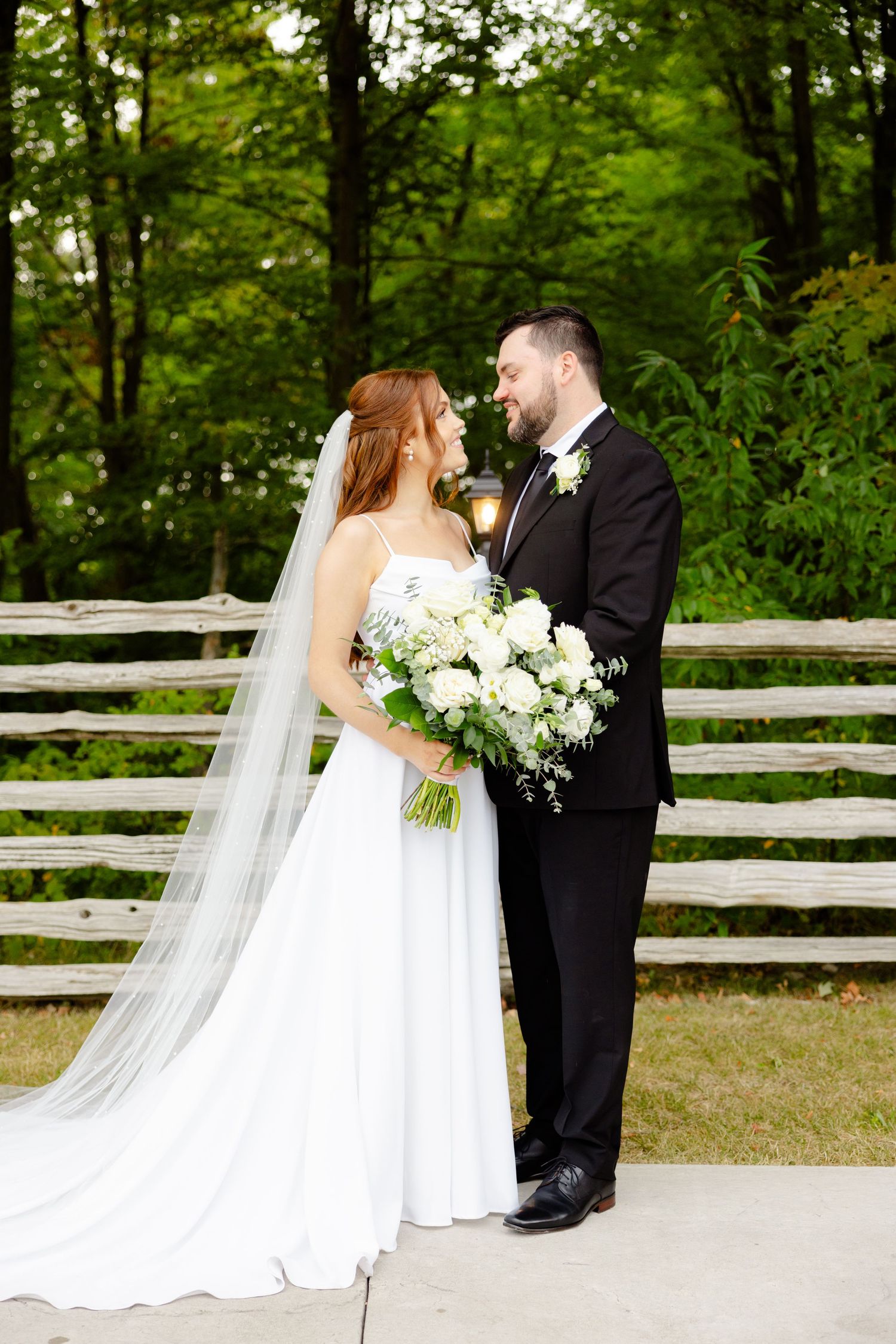 A wedding couple shares a loving moment in front of a rustic wooden fence surrounded by lush green forest.