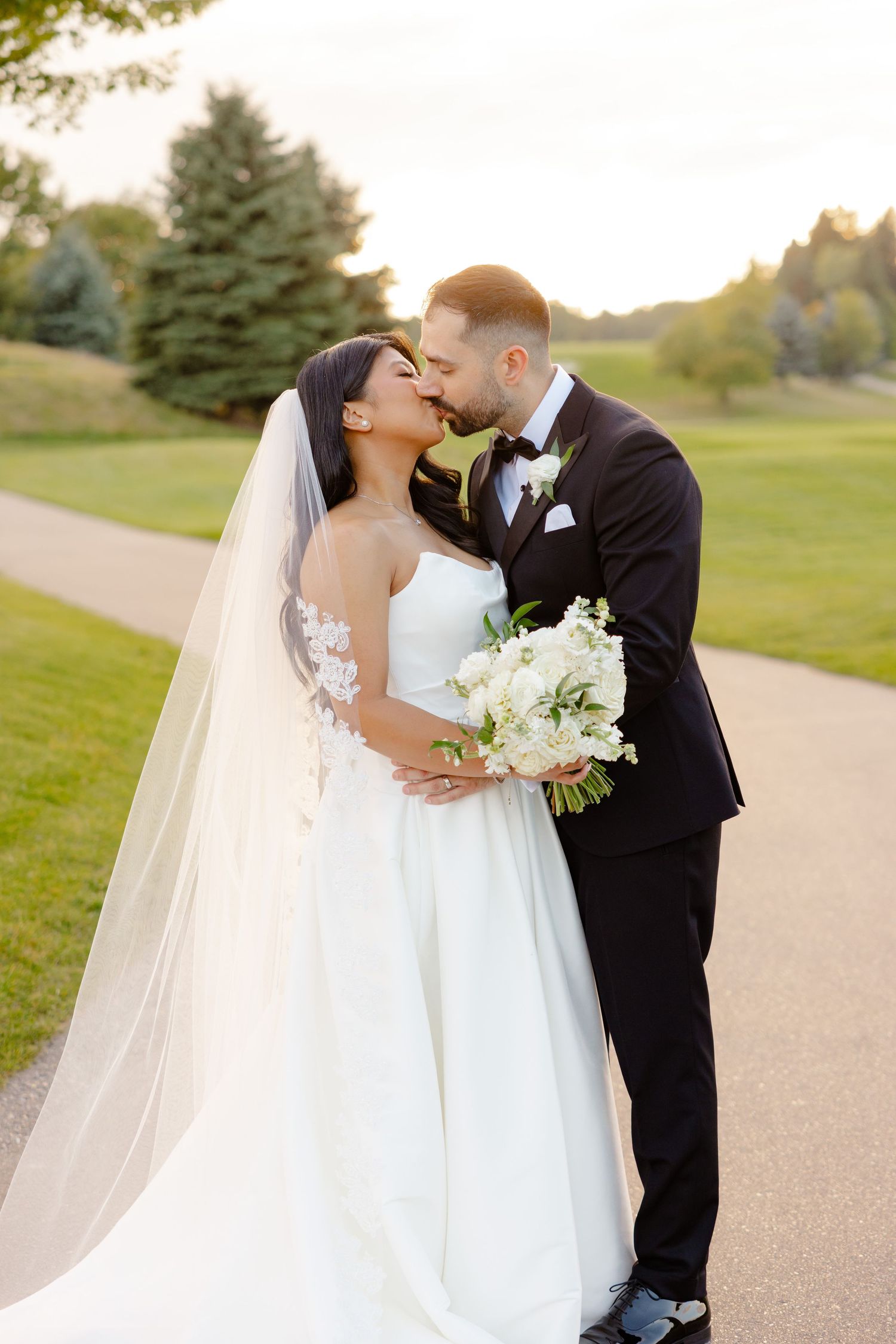 A romantic wedding kiss captured on a scenic golf course path at sunset.