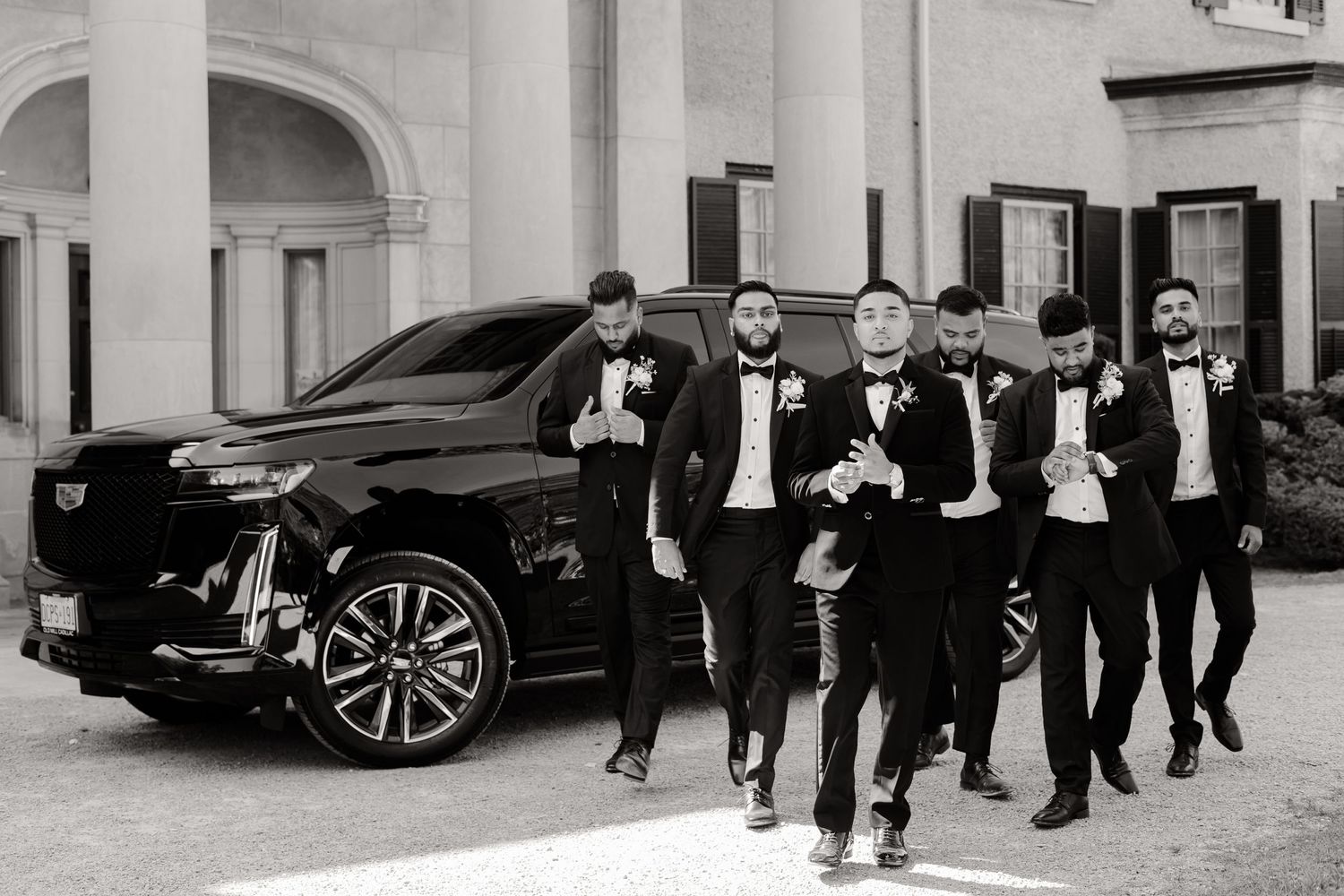 Groomsmen in tuxedos walk together in front of an elegant building and luxury car.