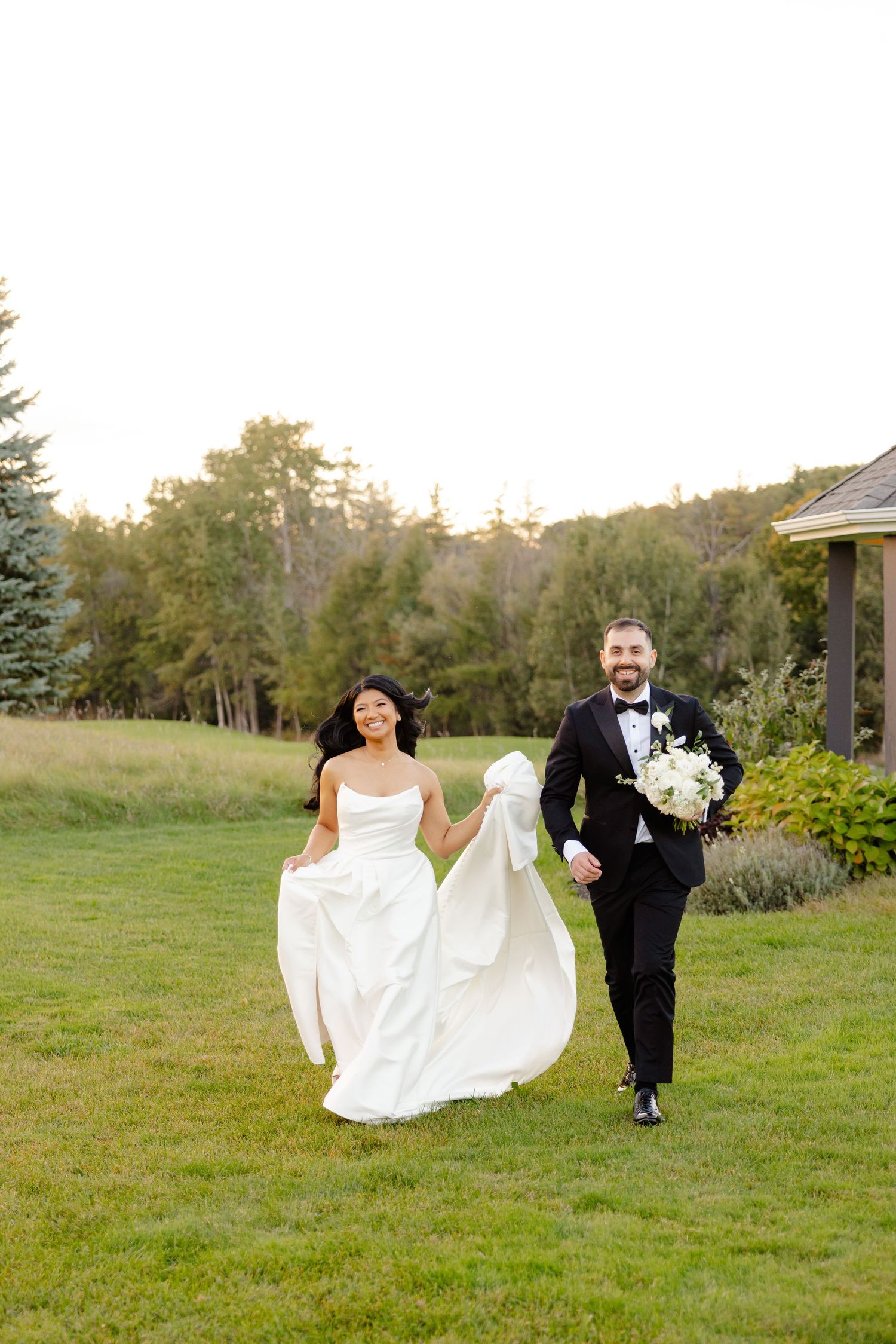 Newlyweds running joyfully across a green lawn with white flowers and garden gazebo in background.