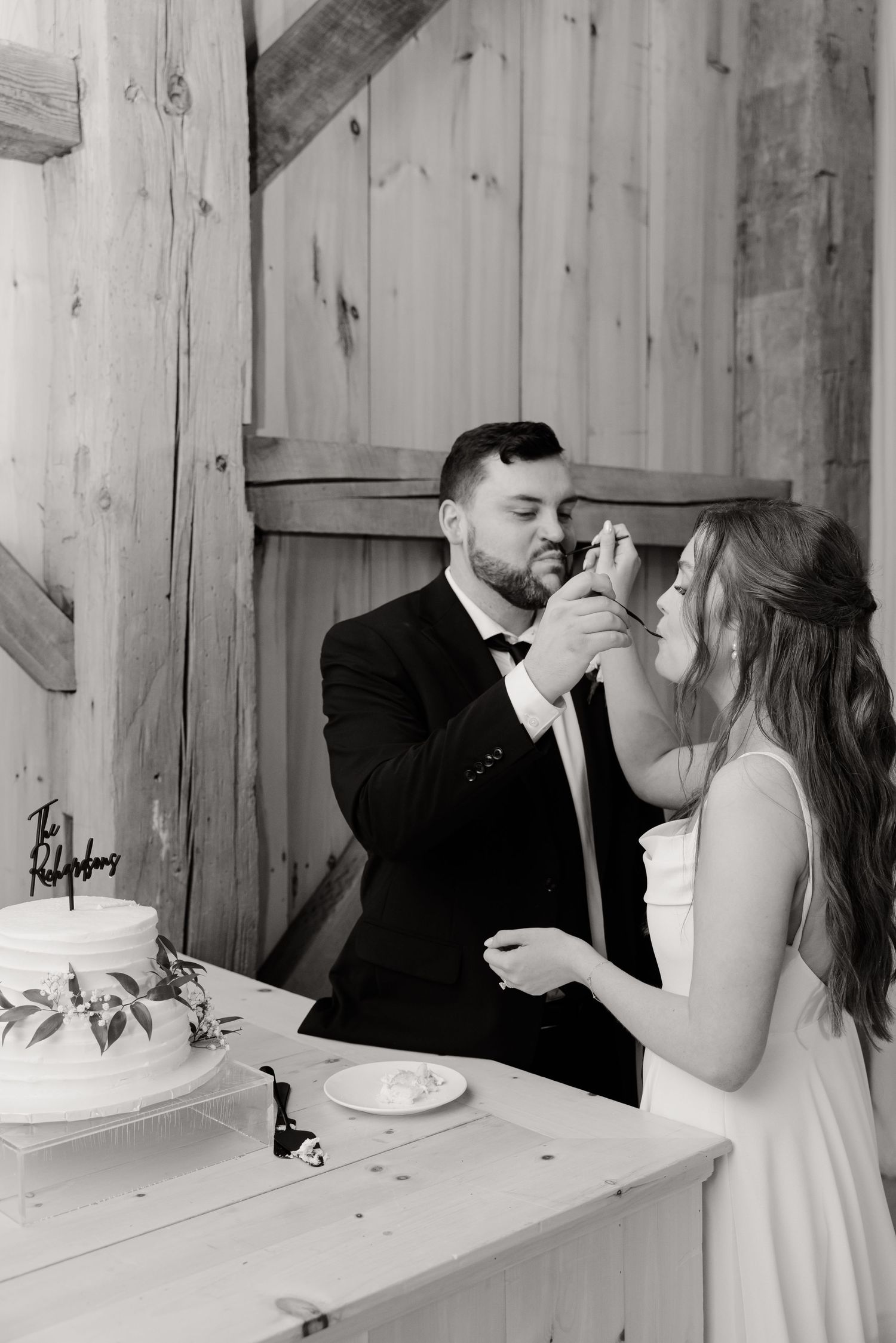 A couple shares cake during their wedding celebration in a rustic wooden barn setting.