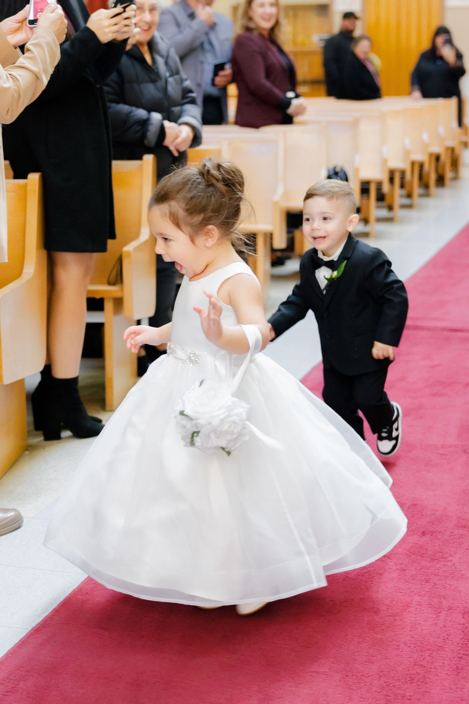 Children in formal attire playfully walk down a red carpet aisle in a church.