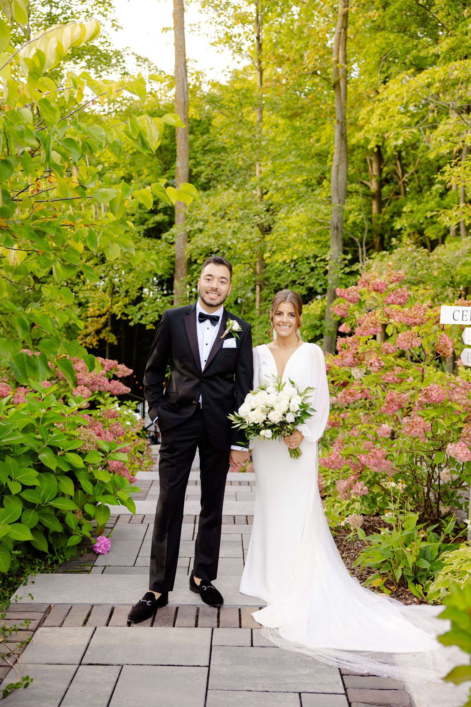 Wedding couple poses on wooden walkway surrounded by vibrant garden flowers and greenery.