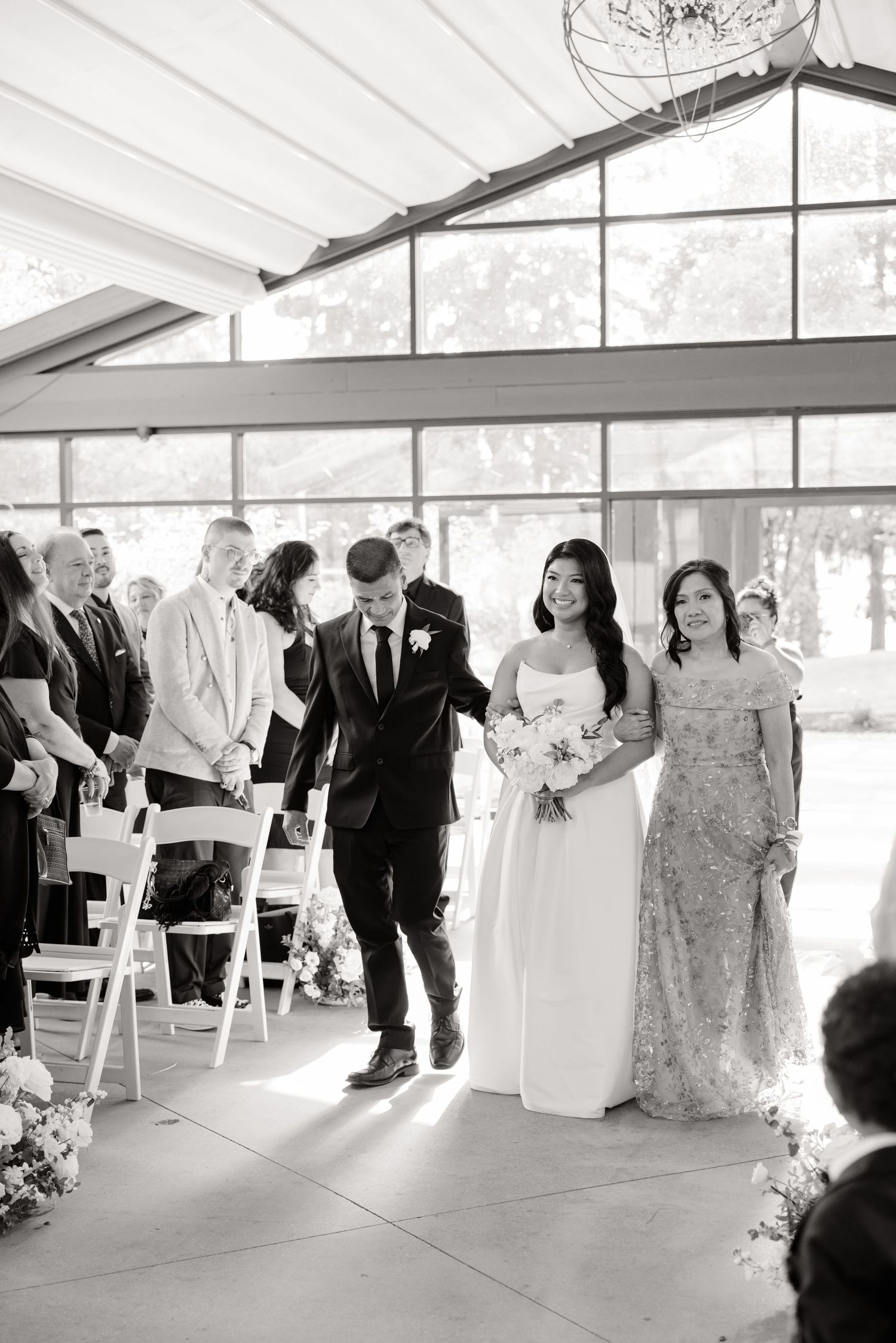 A wedding ceremony processional in a bright indoor venue with white chairs and floral decorations.