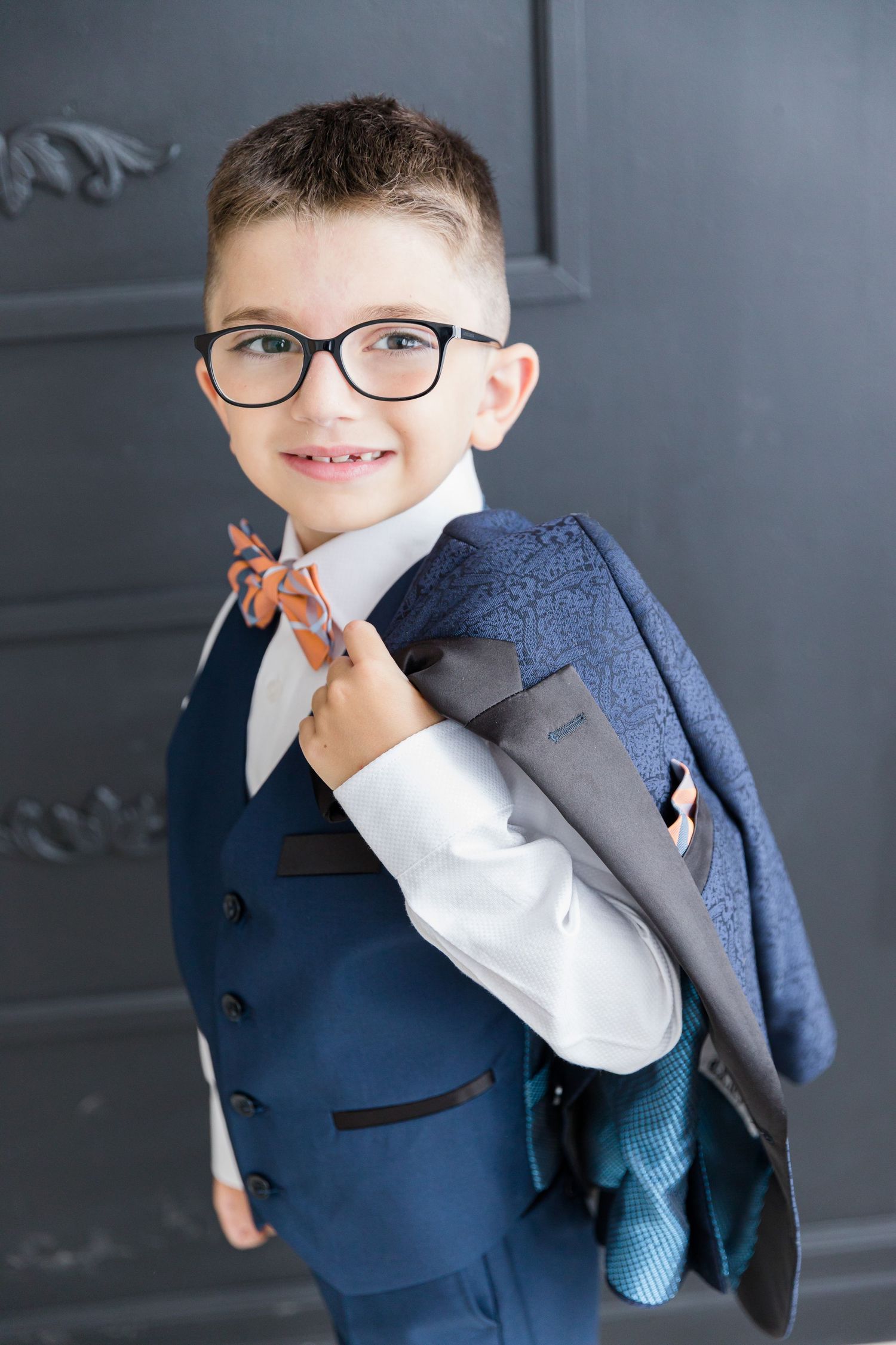 Young boy in navy suit and bow tie holds jacket over shoulder against gray wall.