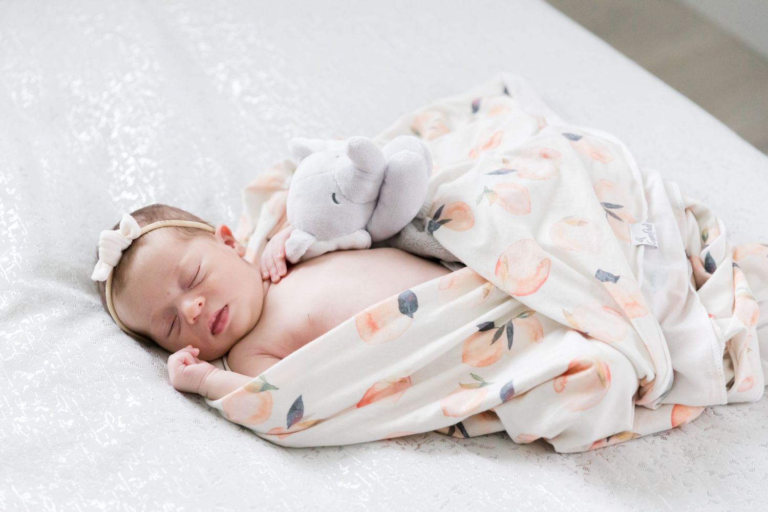 Sleeping newborn wrapped in delicate floral swaddle with white bow headband.