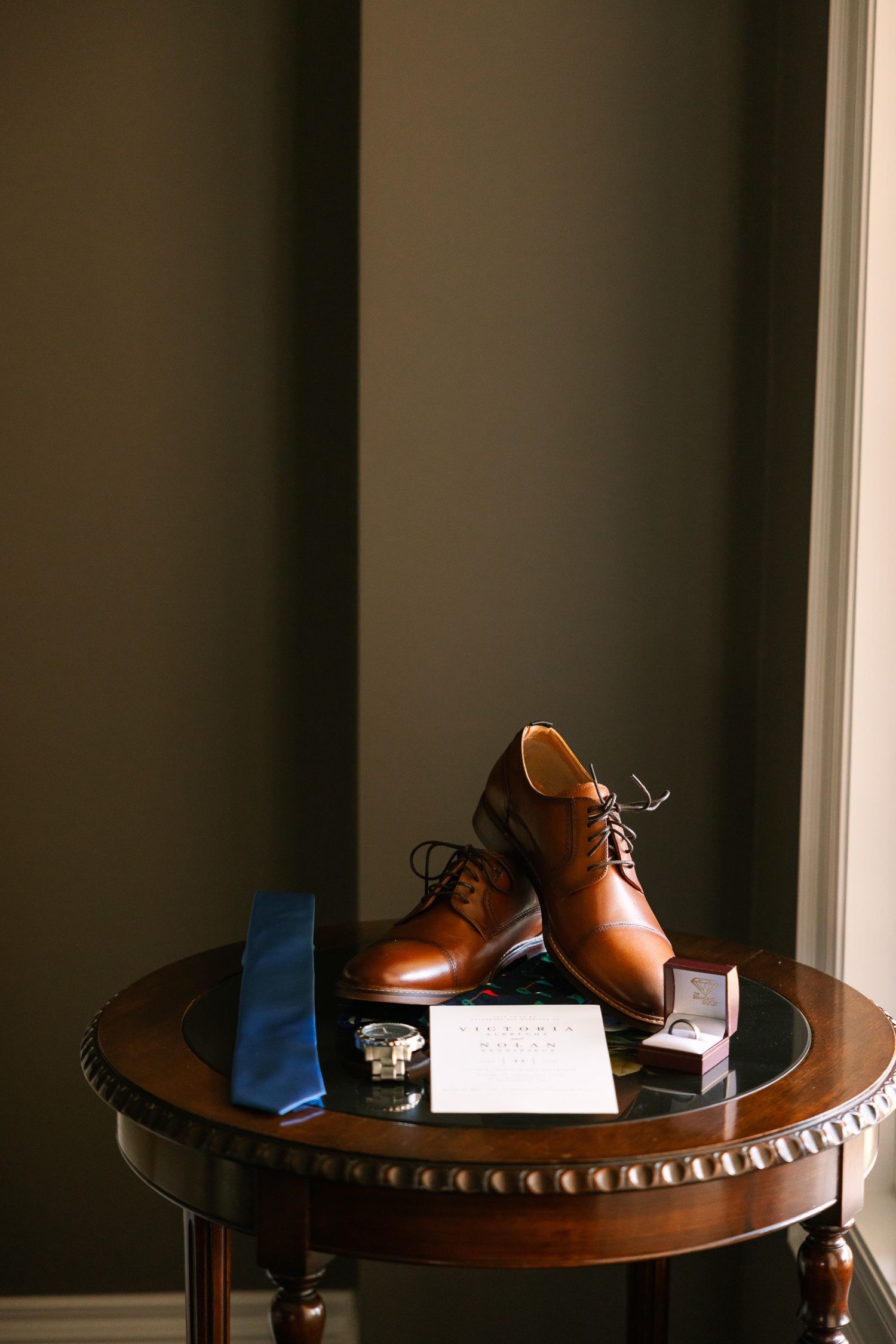 Brown leather dress shoes with blue tie and wedding stationery arranged on wooden table.