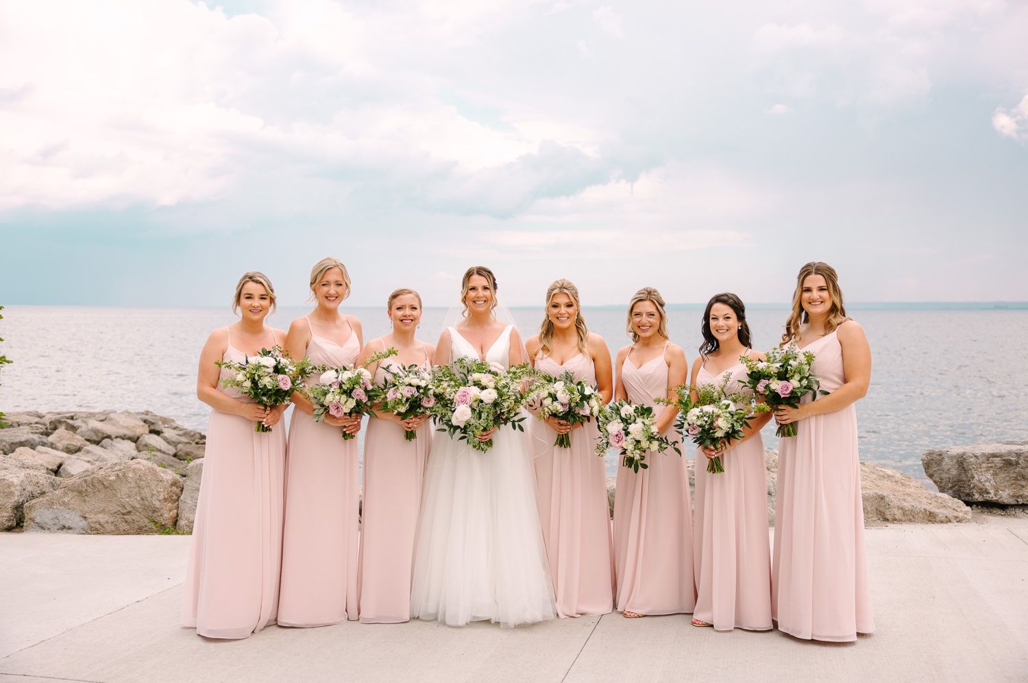 Wedding party wearing pink bridesmaid dresses pose together by the waterfront.