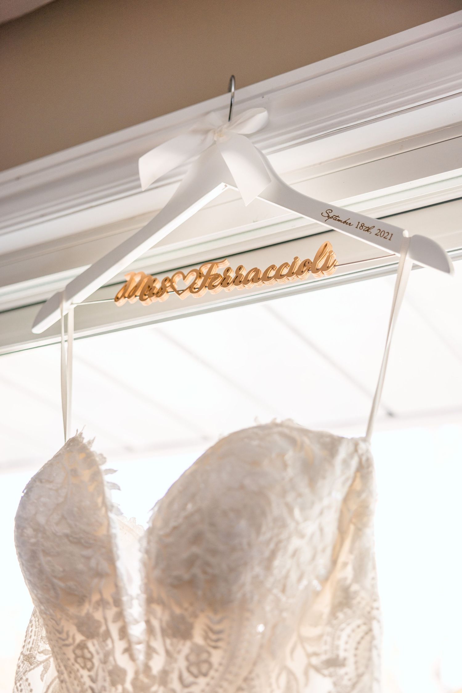 A white satin wedding dress hanger with gold cursive personalization displayed against crown molding.