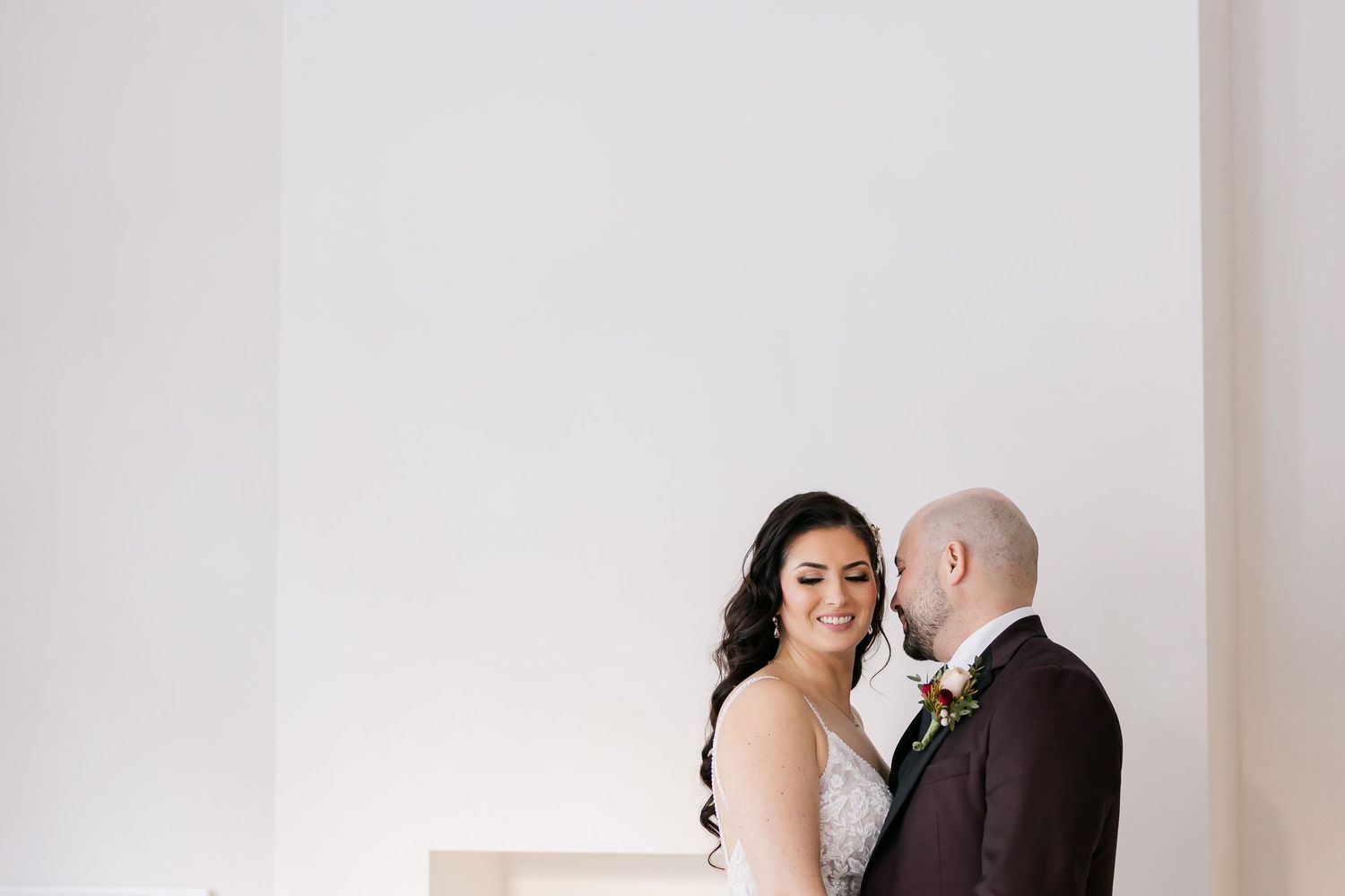A romantic wedding portrait of a couple sharing an intimate moment against a minimalist white wall.