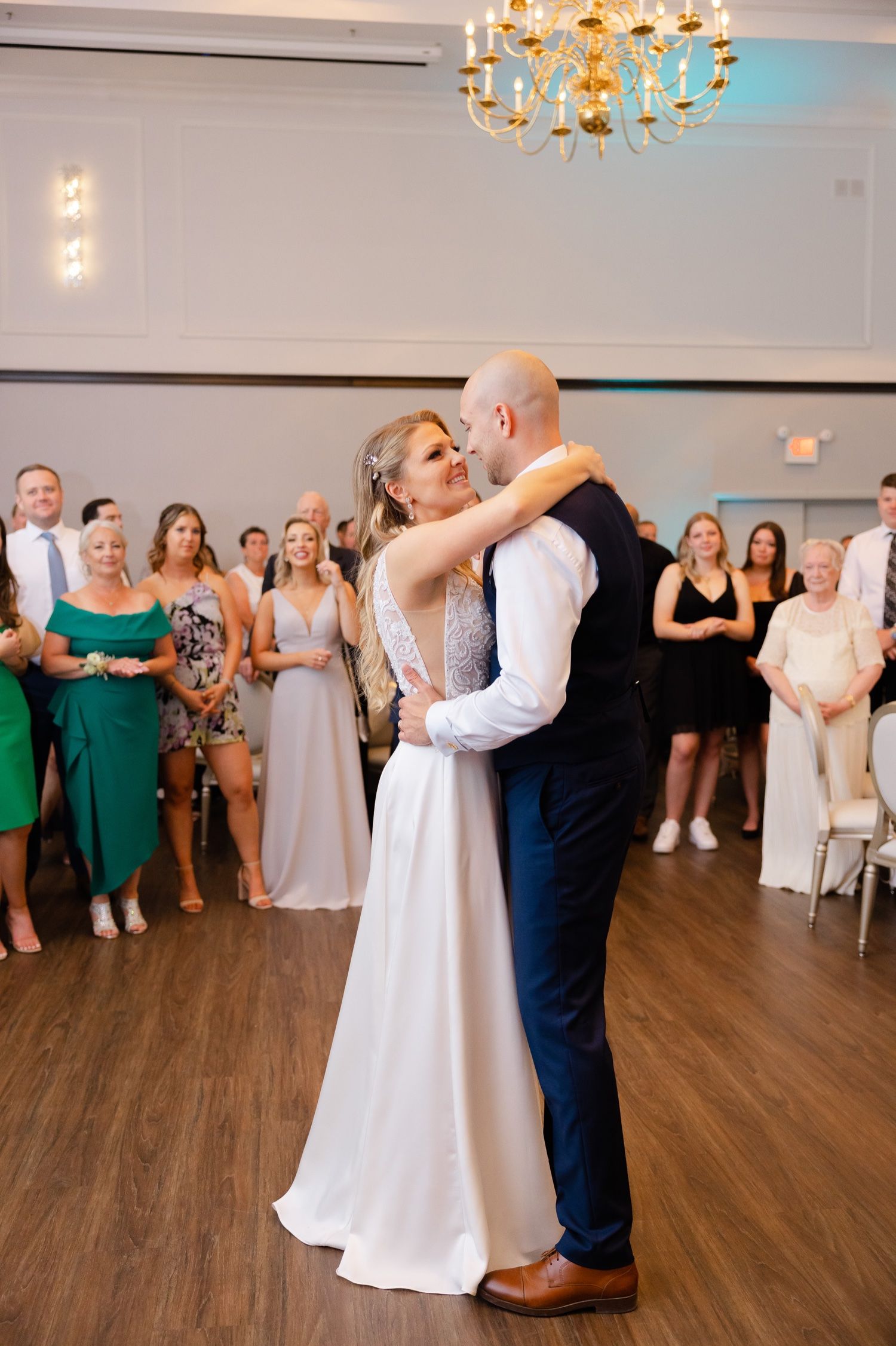 Wedding couple dances while surrounded by guests in elegant reception hall with chandelier.