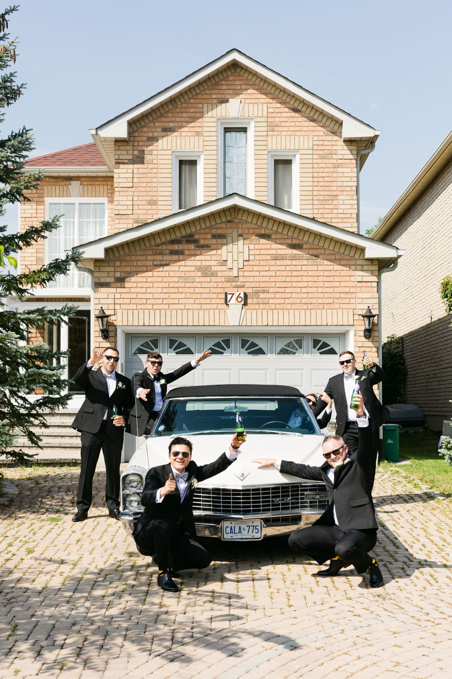 Group in formal attire posing playfully around vintage white car in front of brick house.