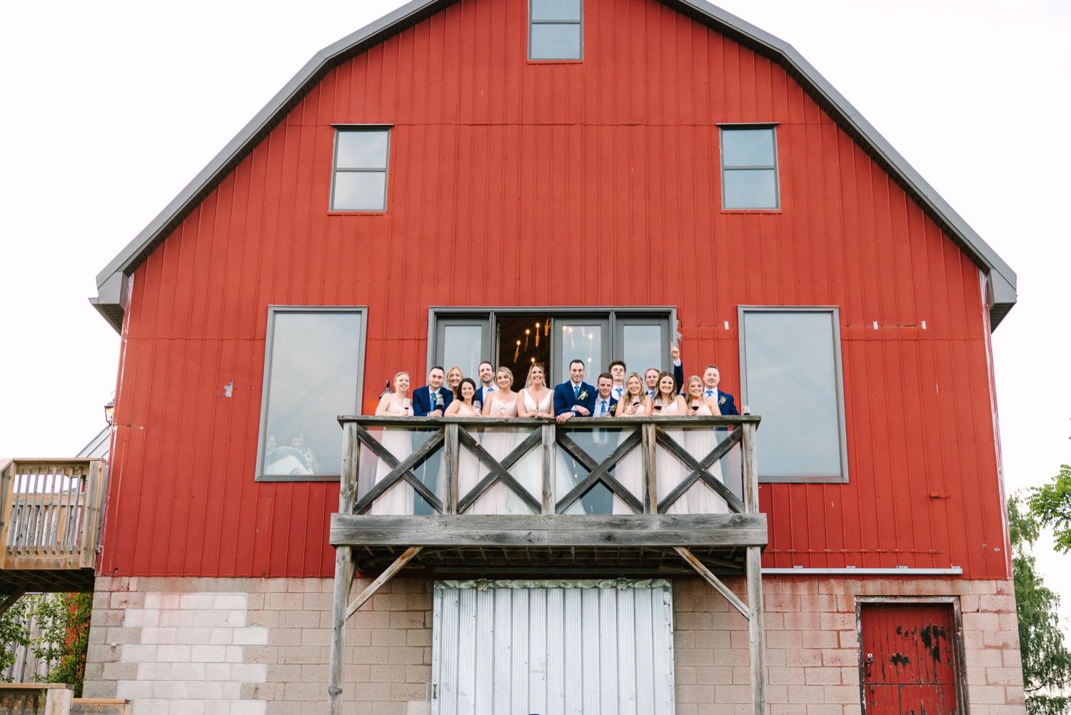 A wedding party poses on the balcony of a vibrant red barn venue.