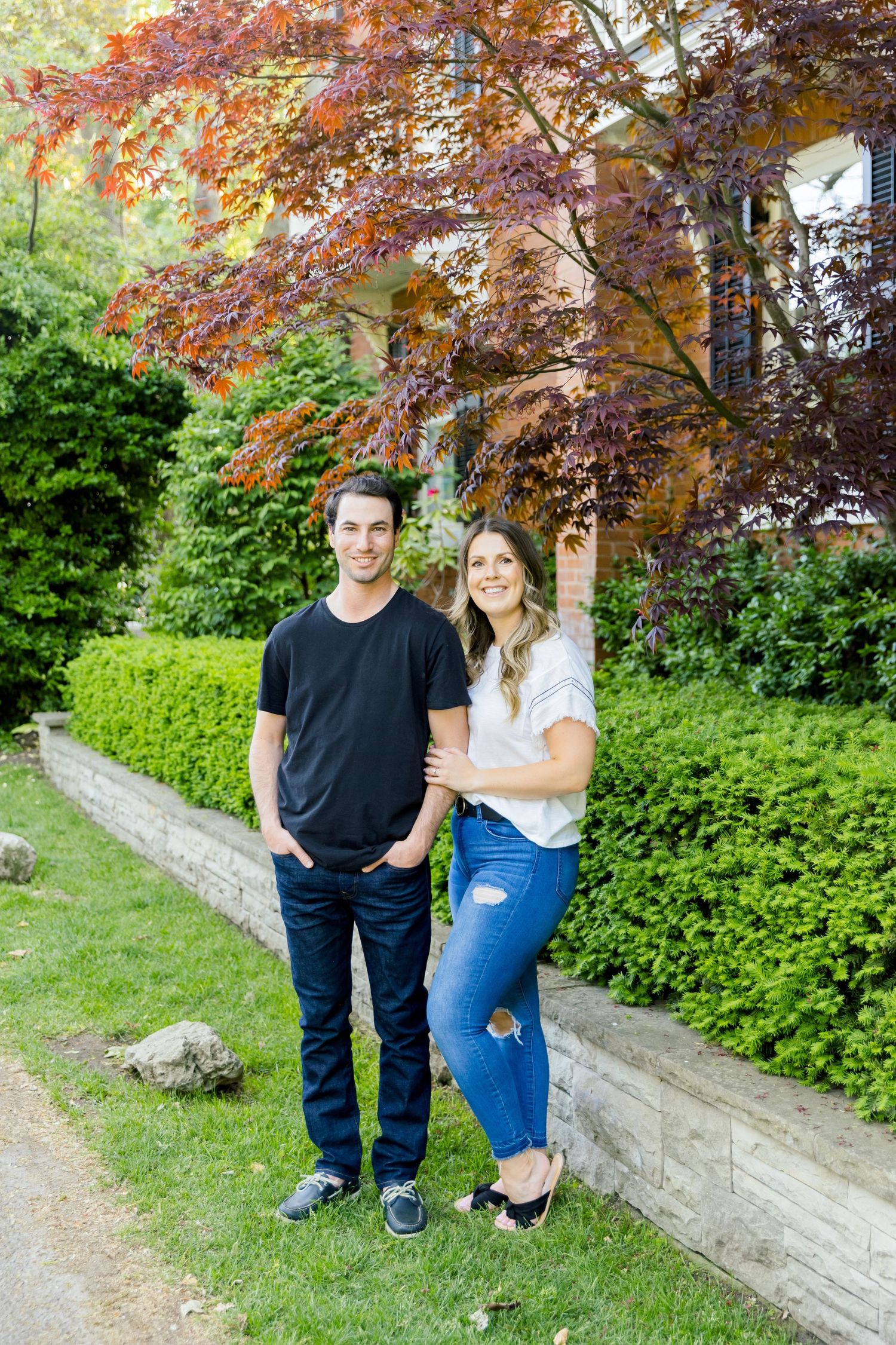 A couple poses for a portrait beneath a Japanese maple tree next to well-maintained hedges.