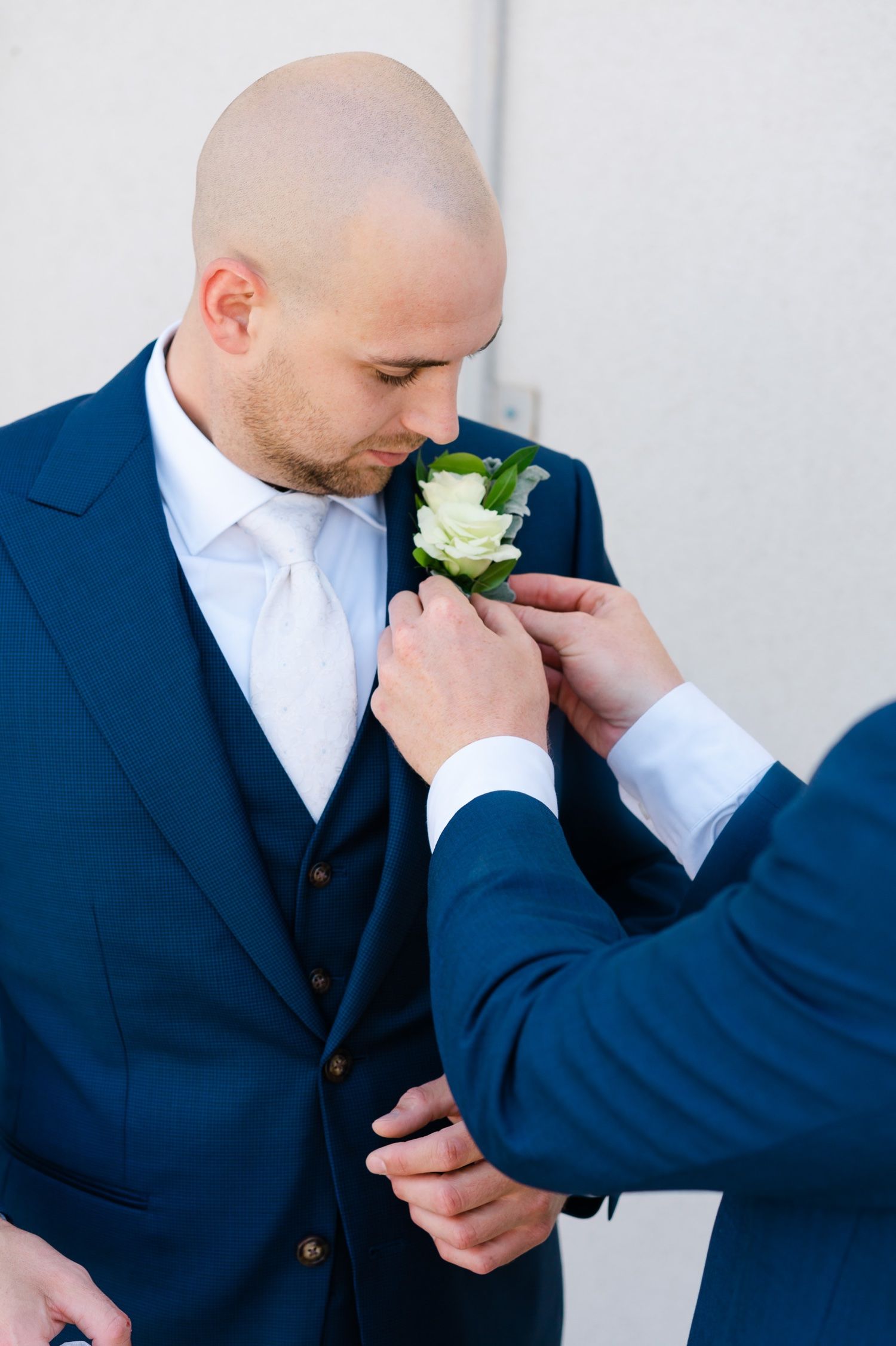 Someone helps pin a white boutonniere flower onto a navy suit lapel.
