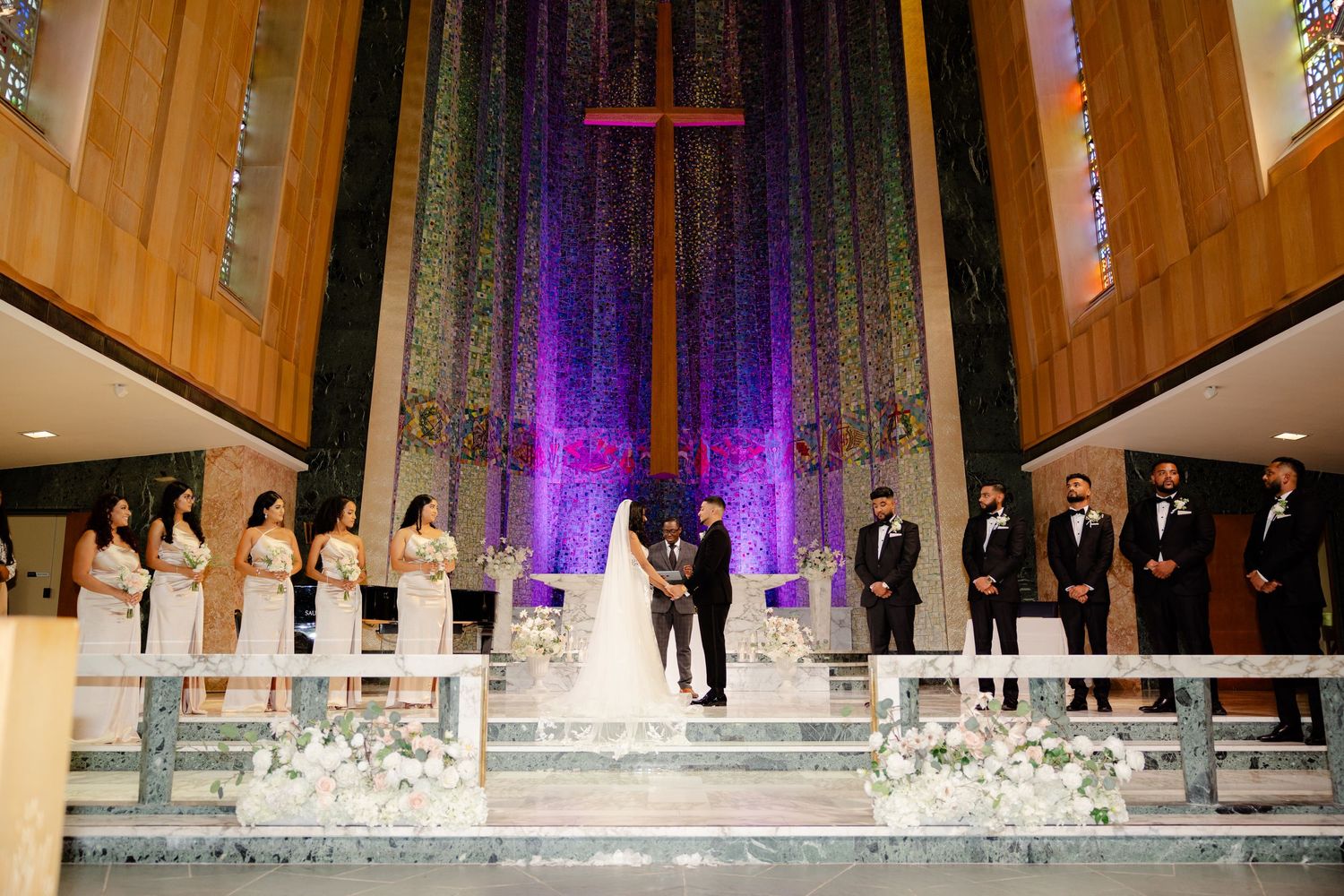 Wedding ceremony in modern church with dramatic purple lighting and white floral arrangements.