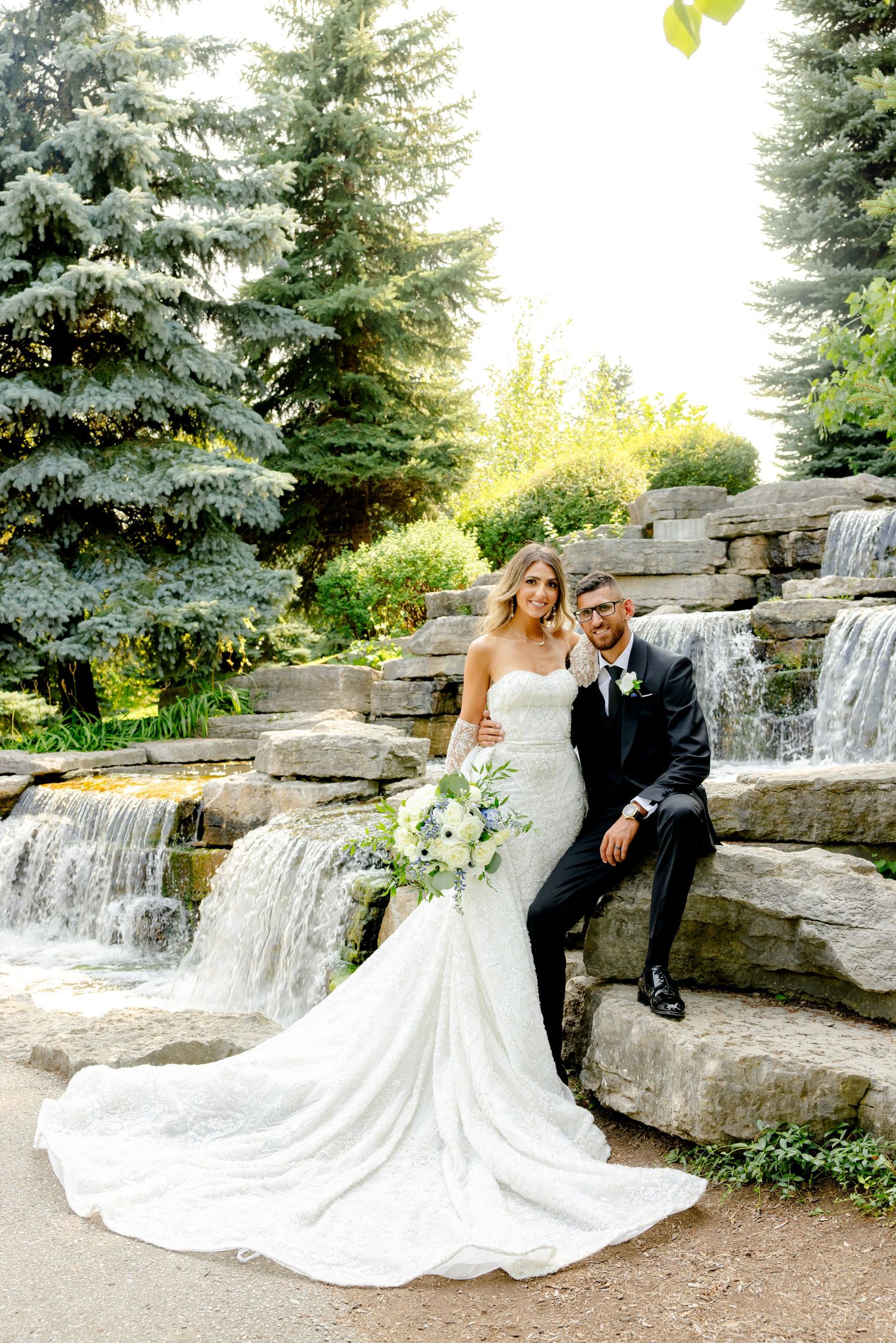 Bride and groom pose together on decorative rock waterfall steps in a garden setting.