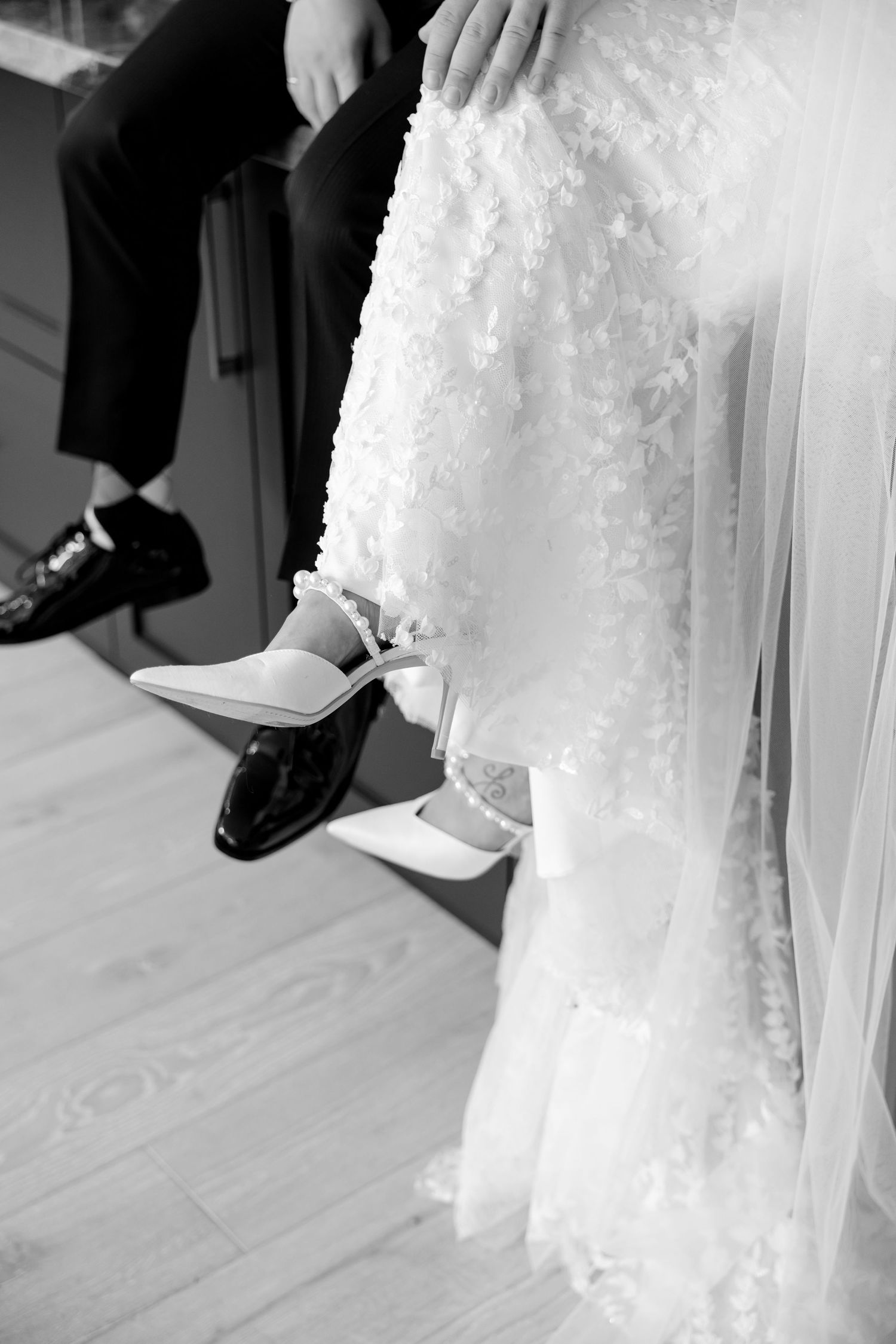 A detail shot of wedding shoes showing black dress shoes and white heels on a light wooden floor.