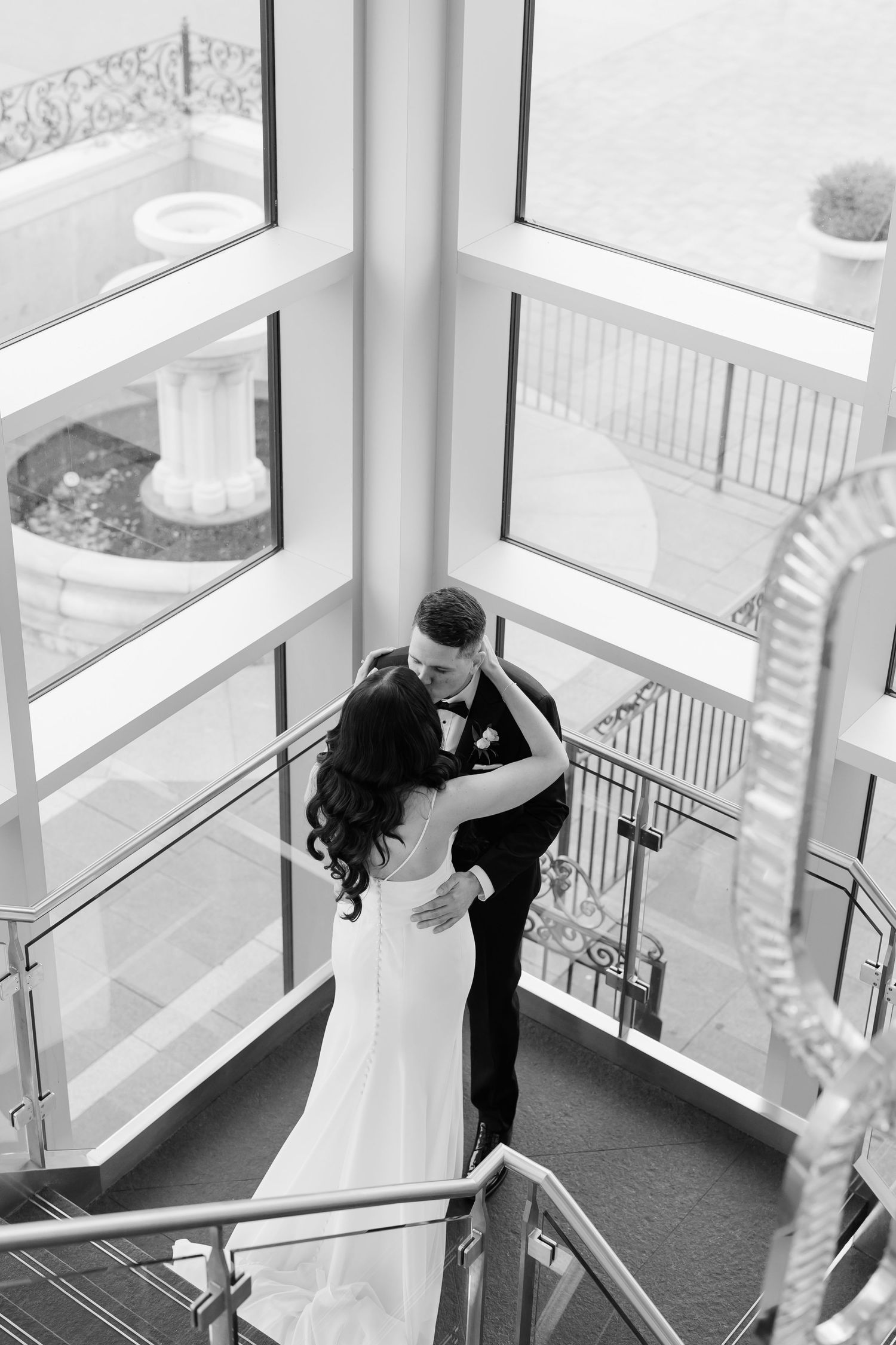 Black and white portrait of newlyweds embracing in an elegant stairwell with floor-to-ceiling windows.