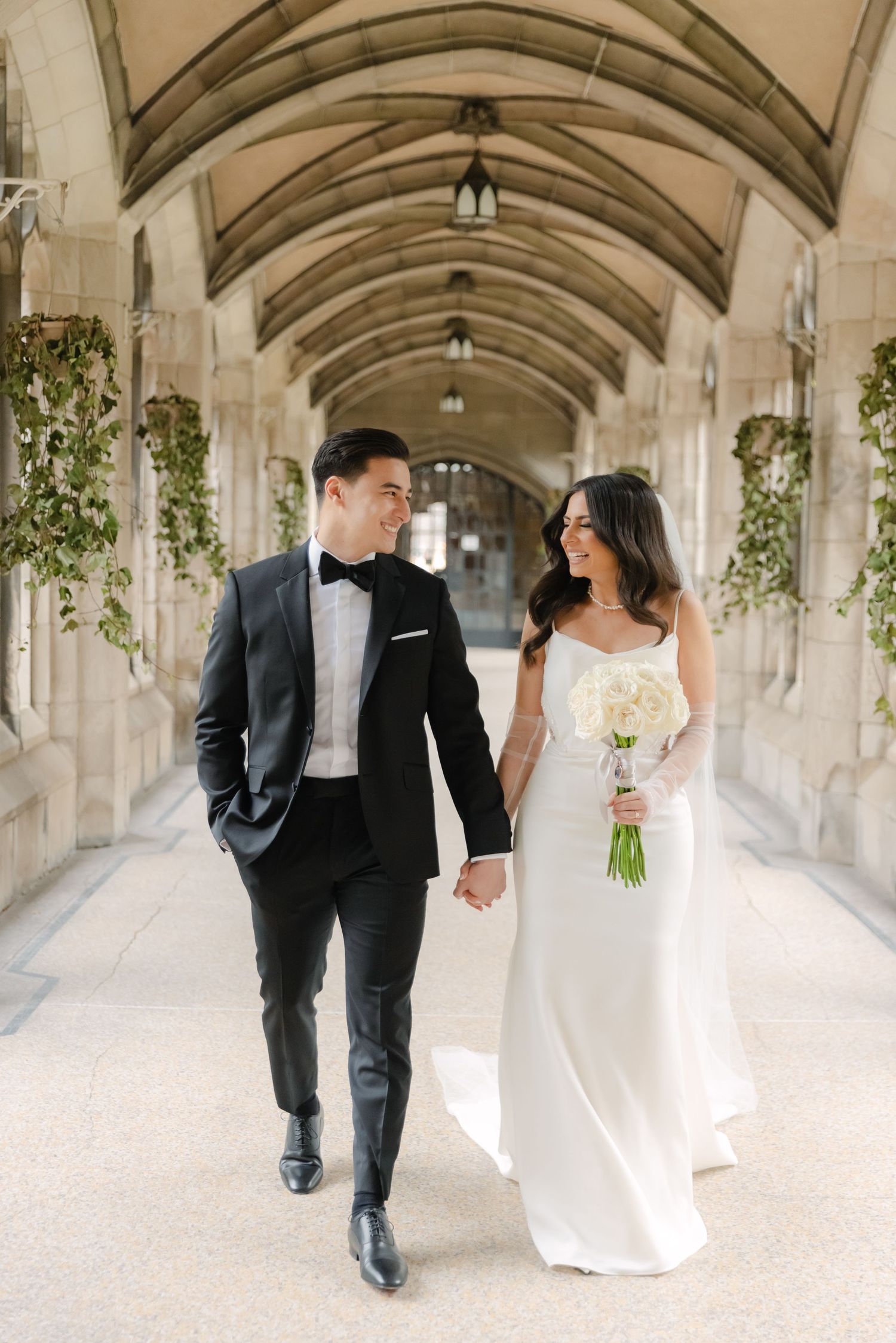 Newlyweds hold hands while walking through a grand stone colonnade with hanging greenery.