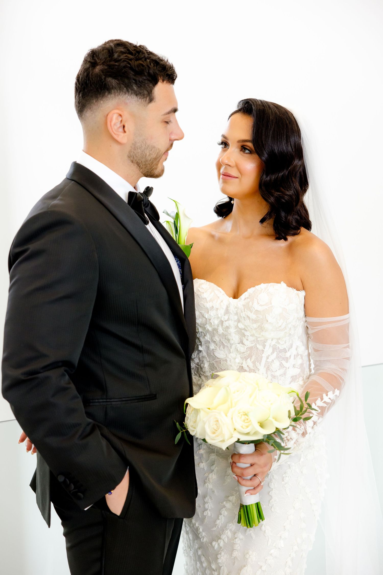 A bride in a beautiful lace gown holds a white bouquet while sharing an intimate moment with her groom in a black tuxedo.