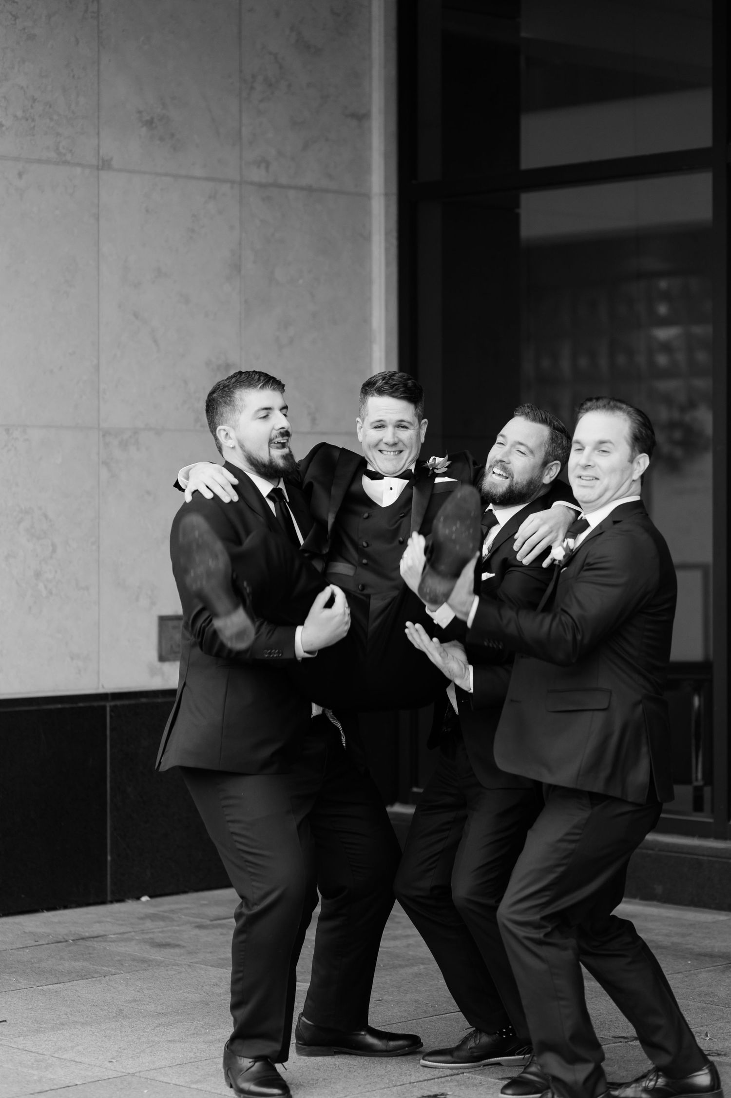 Groomsmen in black suits posing playfully and laughing together in a candid black and white moment.