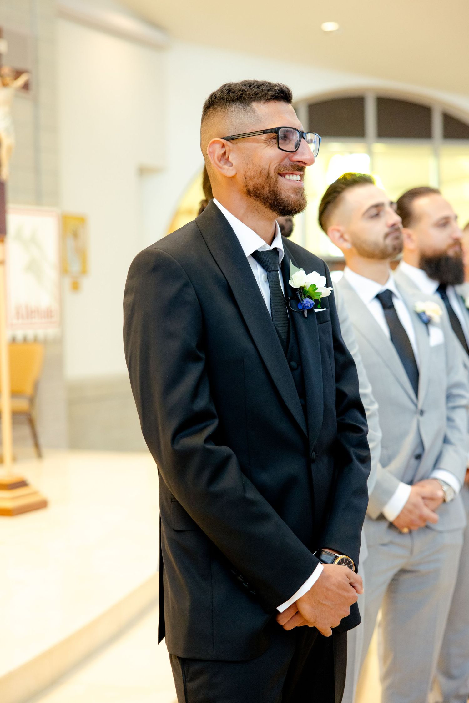 A groomsman in a black suit and tie smiles during a wedding ceremony inside a church.