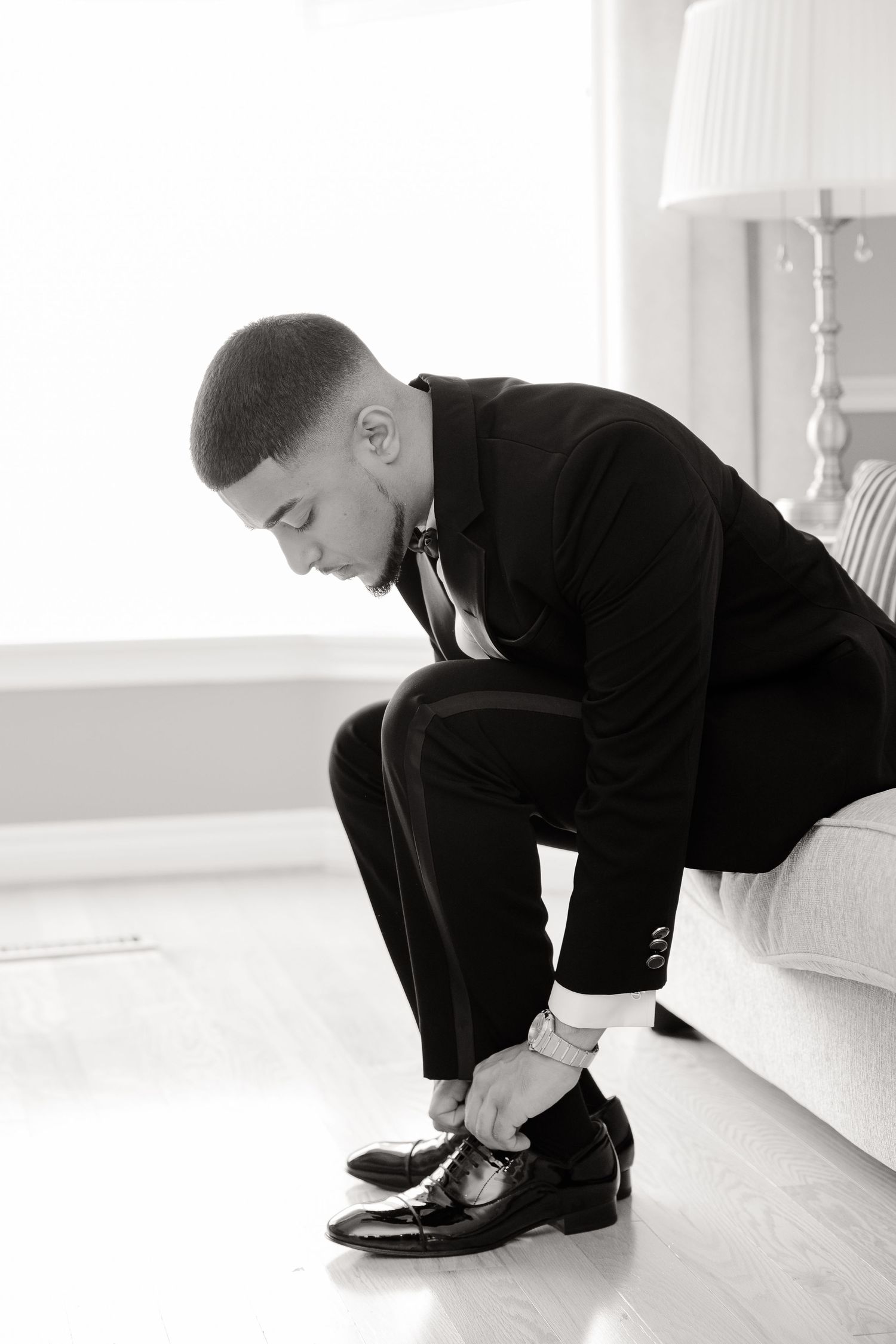 Formal black-tie gettting ready moment showing person tying patent leather dress shoes while sitting on white couch.