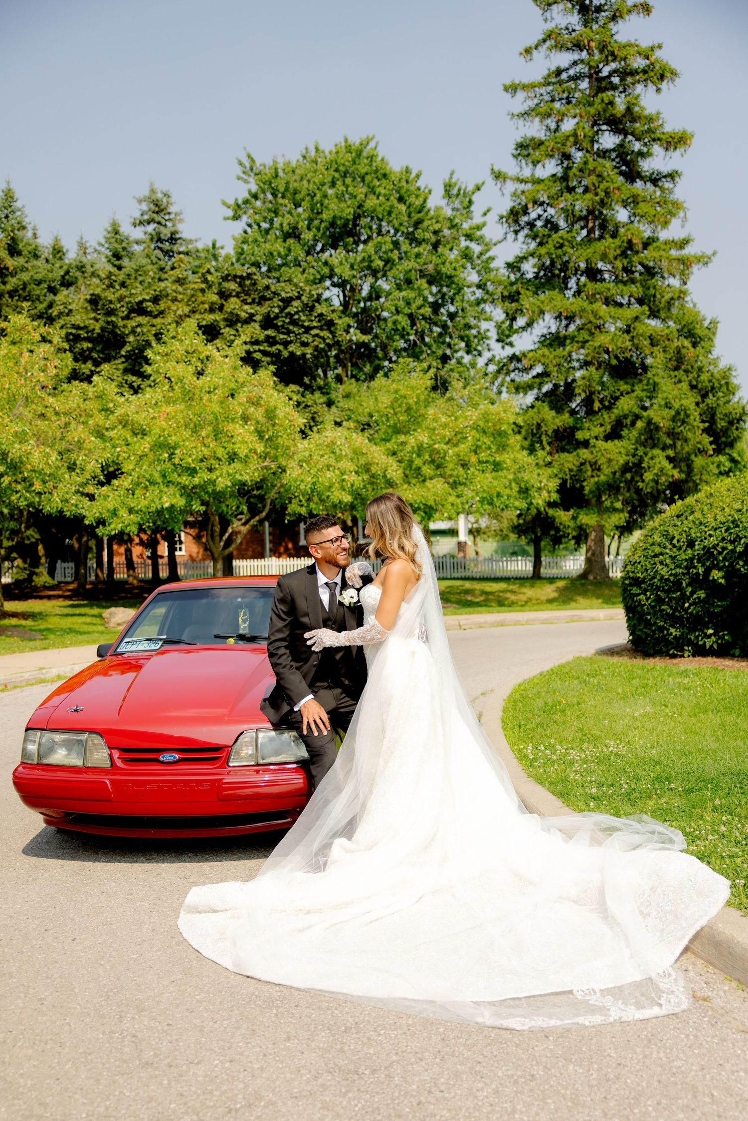 Wedding couple poses with their red car against a backdrop of lush green trees.