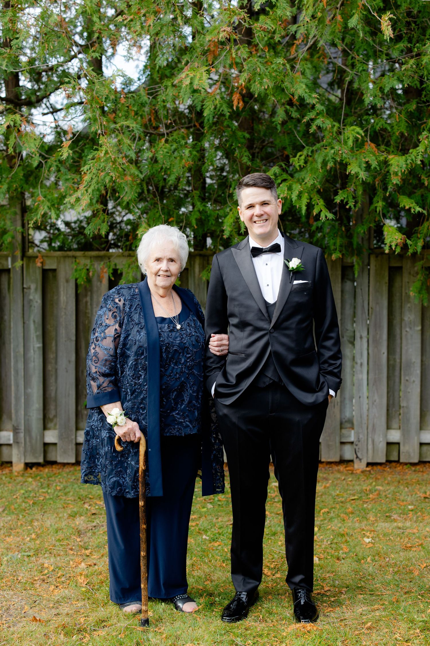 A formal portrait of two people in navy and black formal wear standing against a wooden fence backdrop.