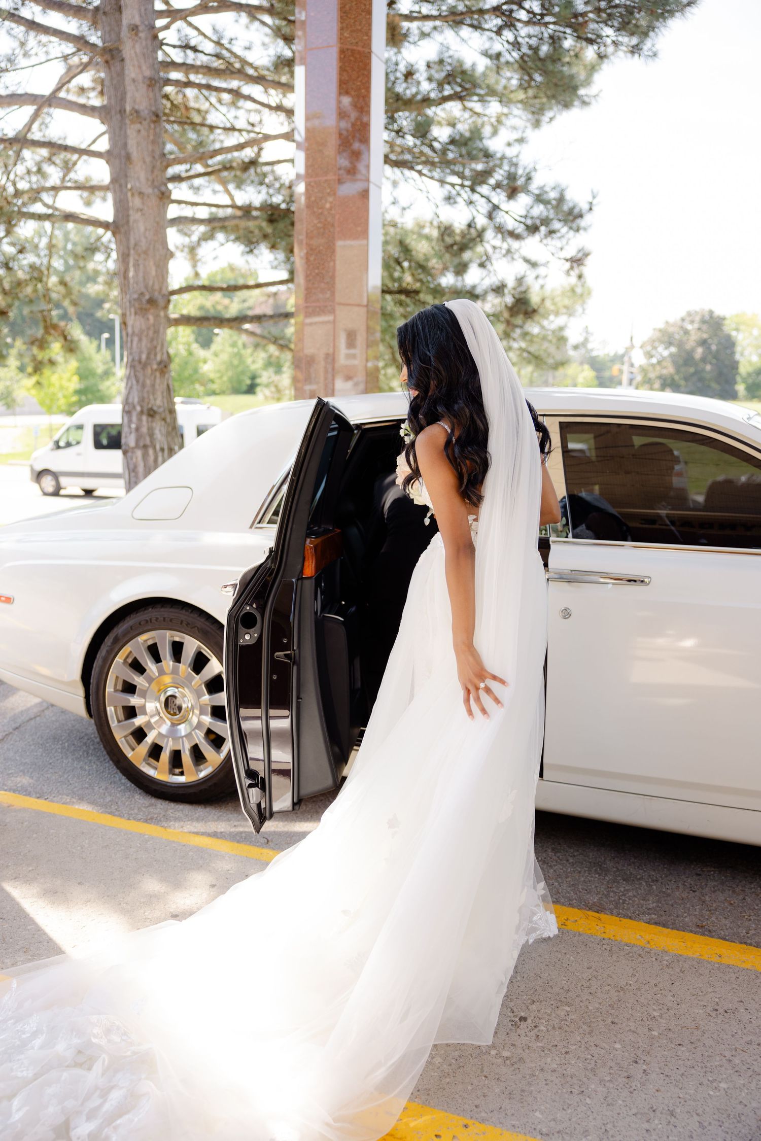 Bride in flowing white gown entering luxury white car after wedding ceremony.
