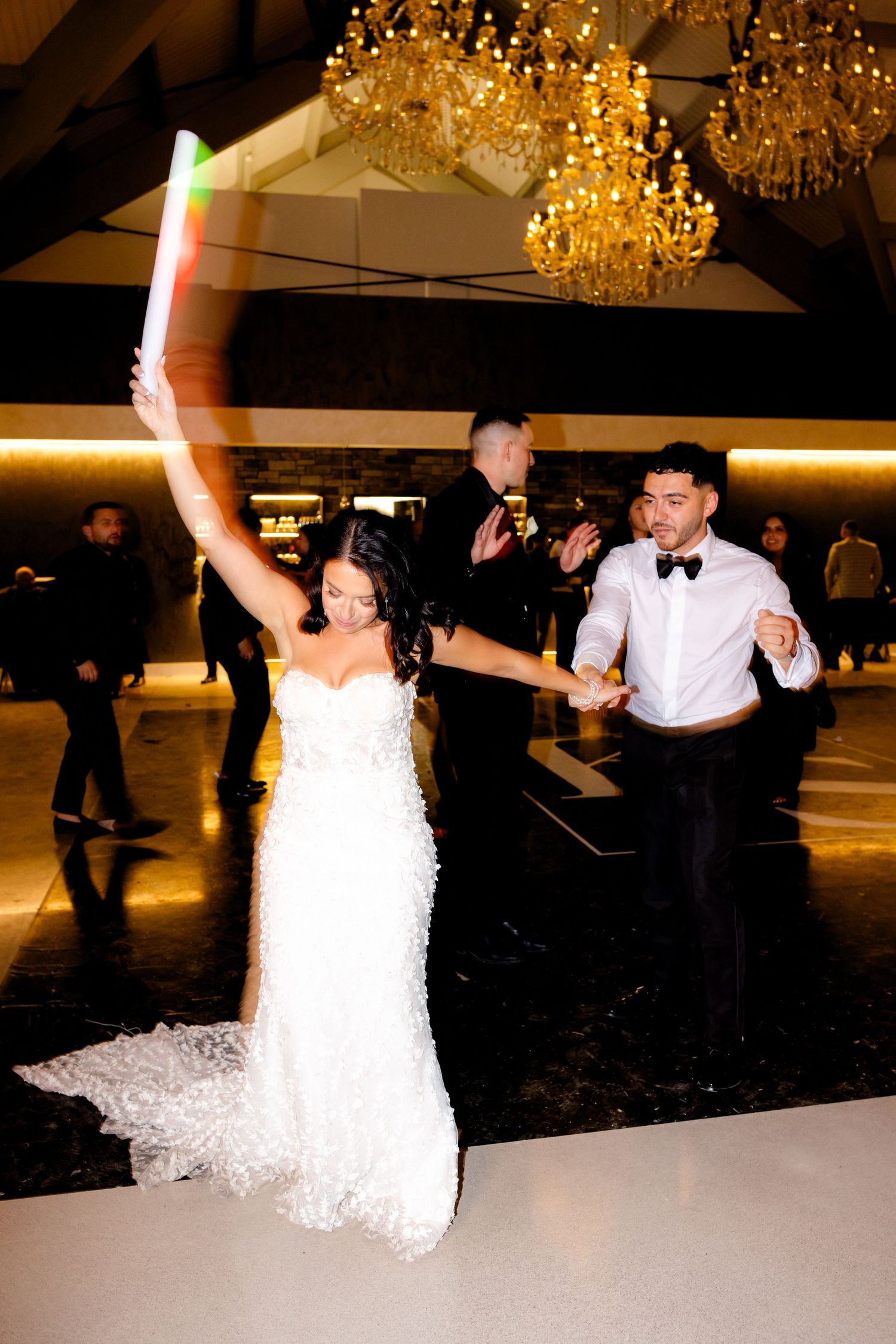 Wedding celebration under golden chandeliers as couple dances energetically on white floor.