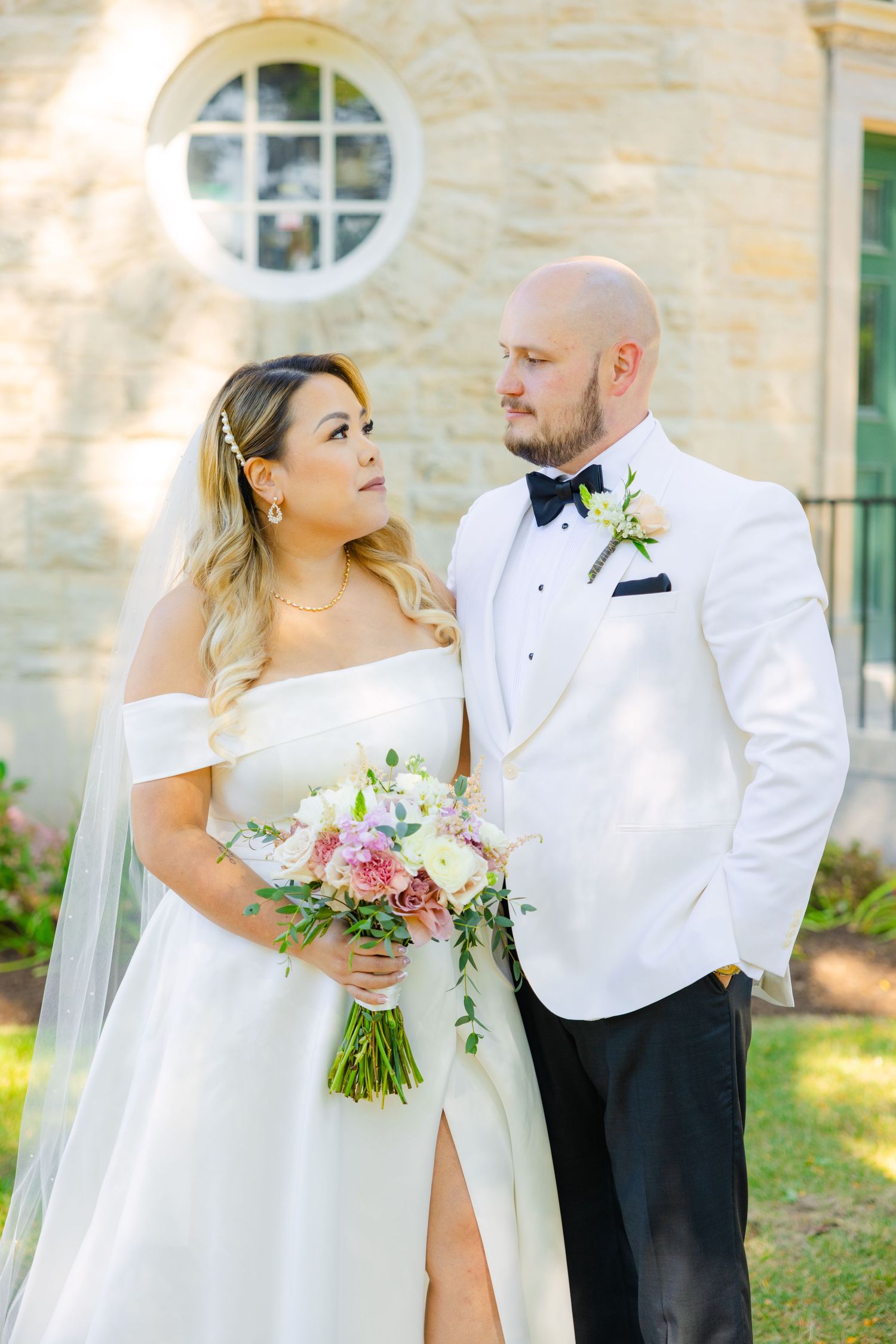 A couple shares a romantic moment in their wedding attire beside a stone building with flowering garden.