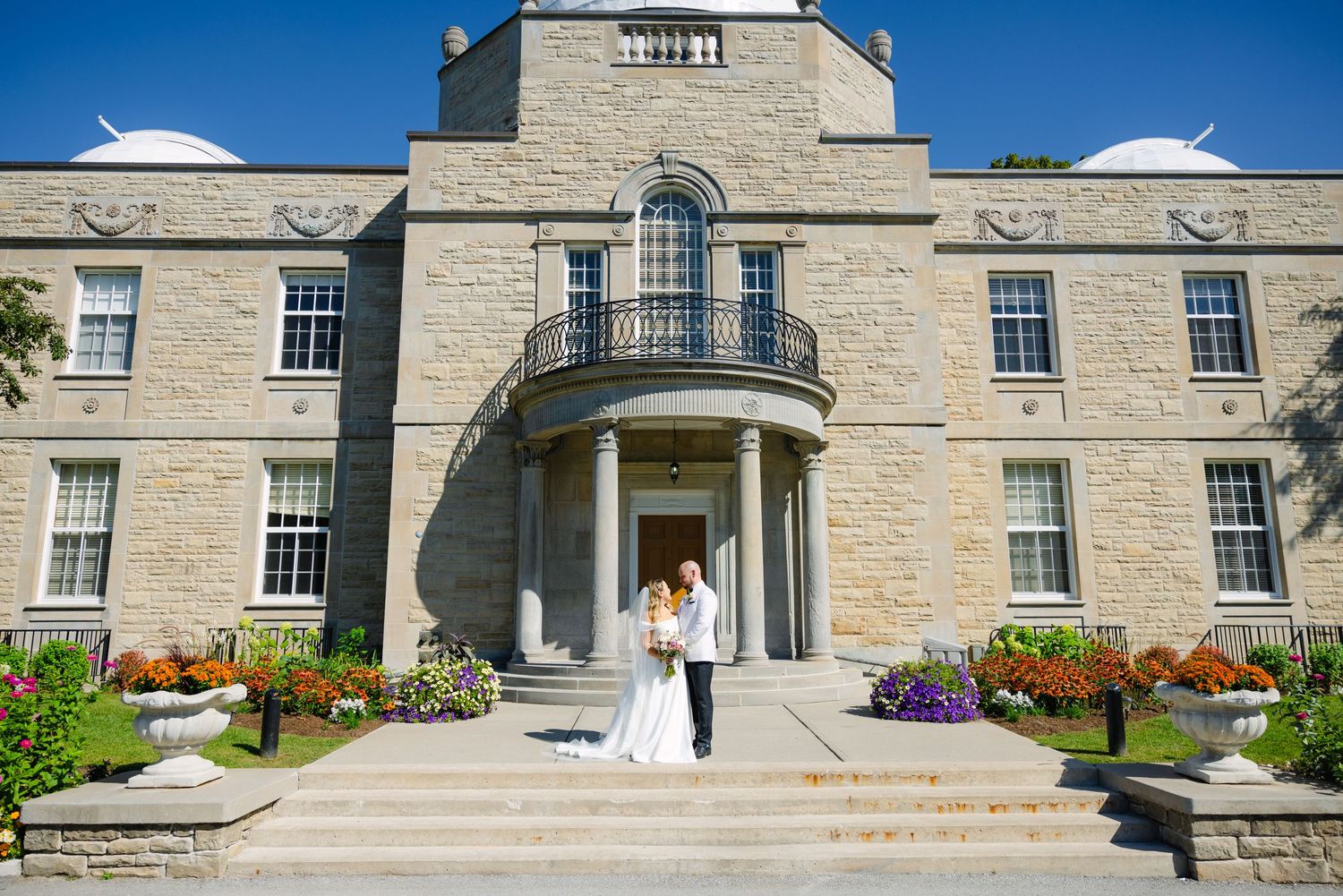 Wedding couple posing in front of a limestone mansion with classic architecture and blooming flowers.