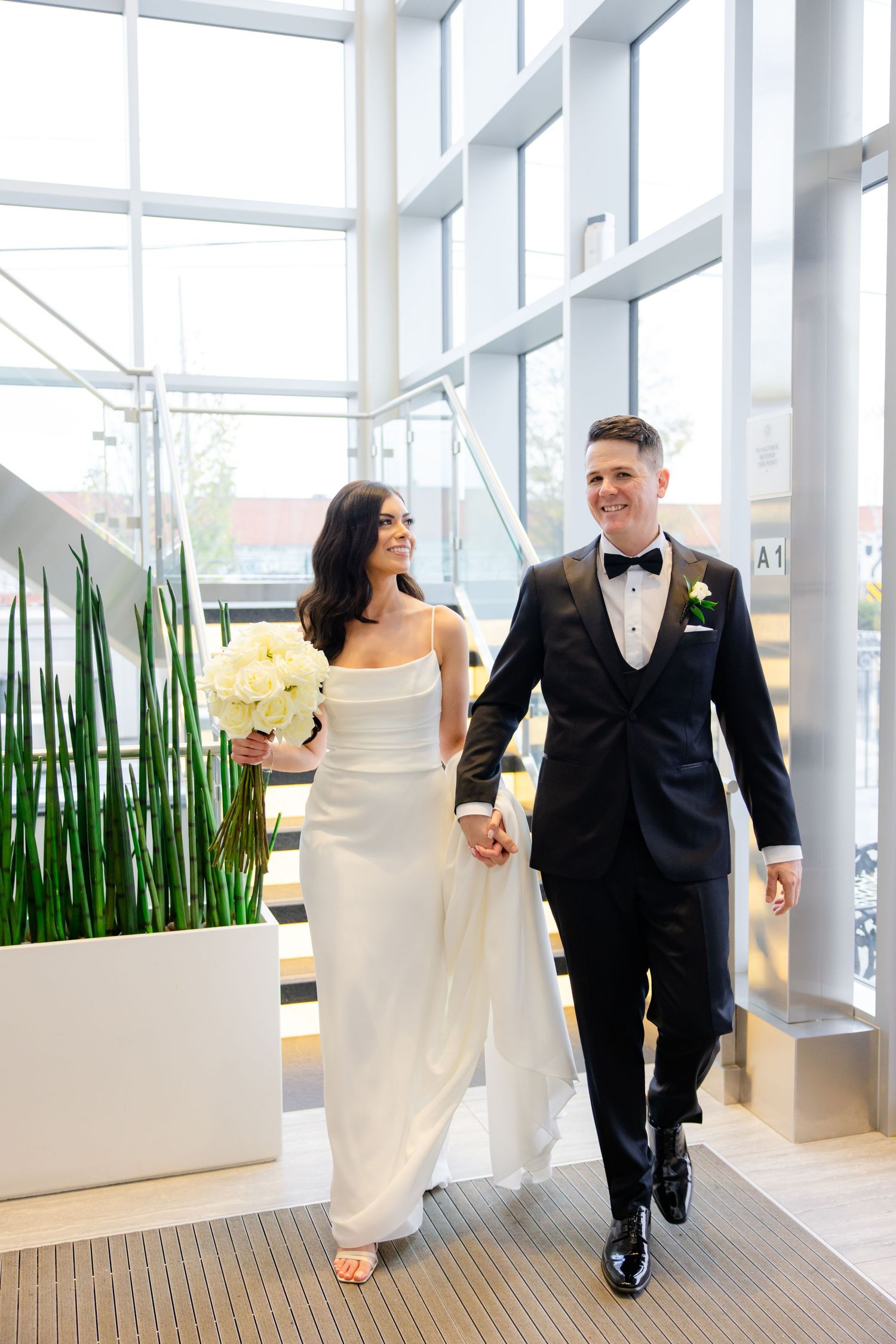 Elegant couple walking through modern venue with floor-to-ceiling windows and indoor plants.