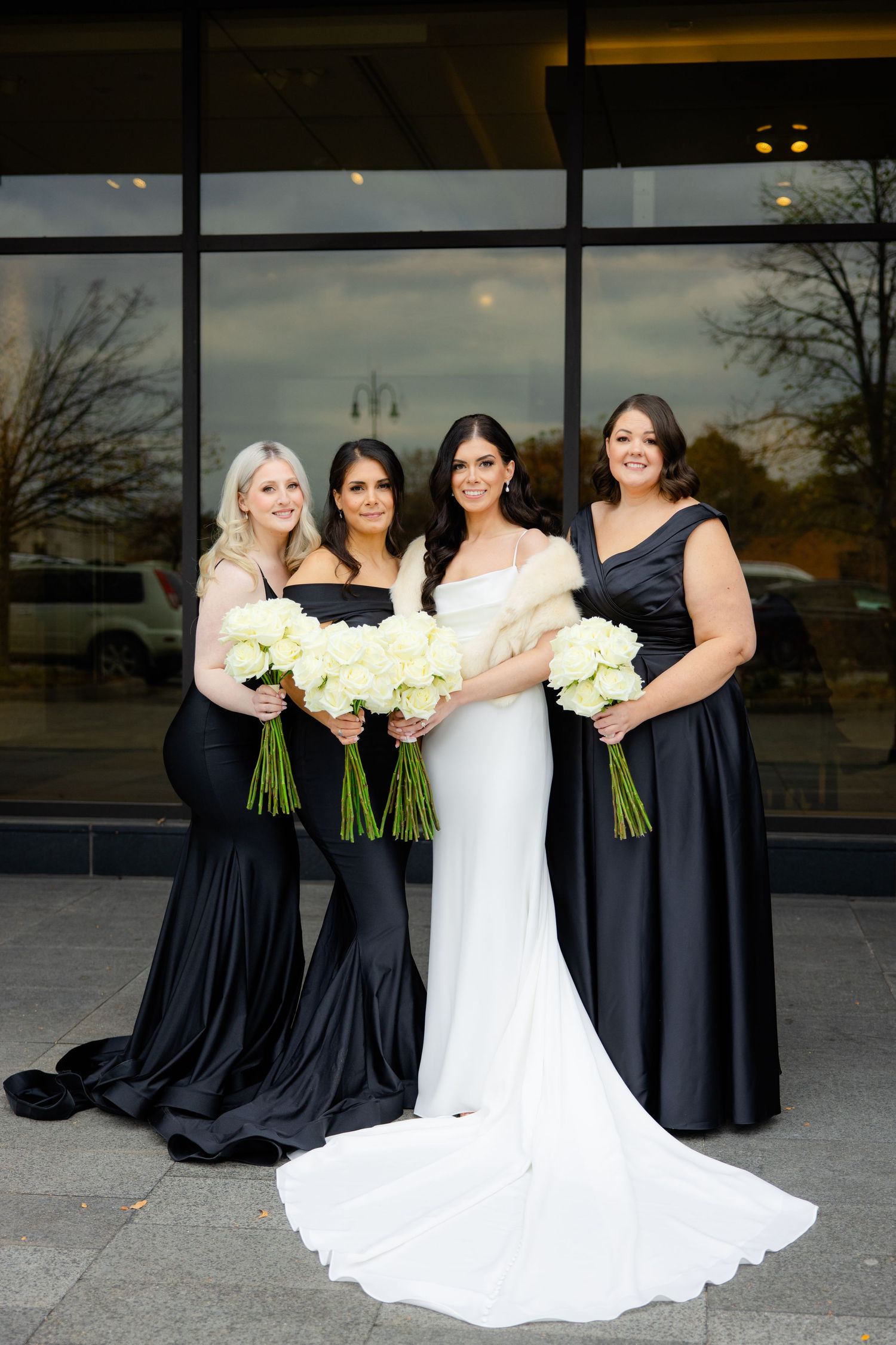 Wedding party poses with bride in white gown and bridesmaids in flowing black dresses holding bouquets.