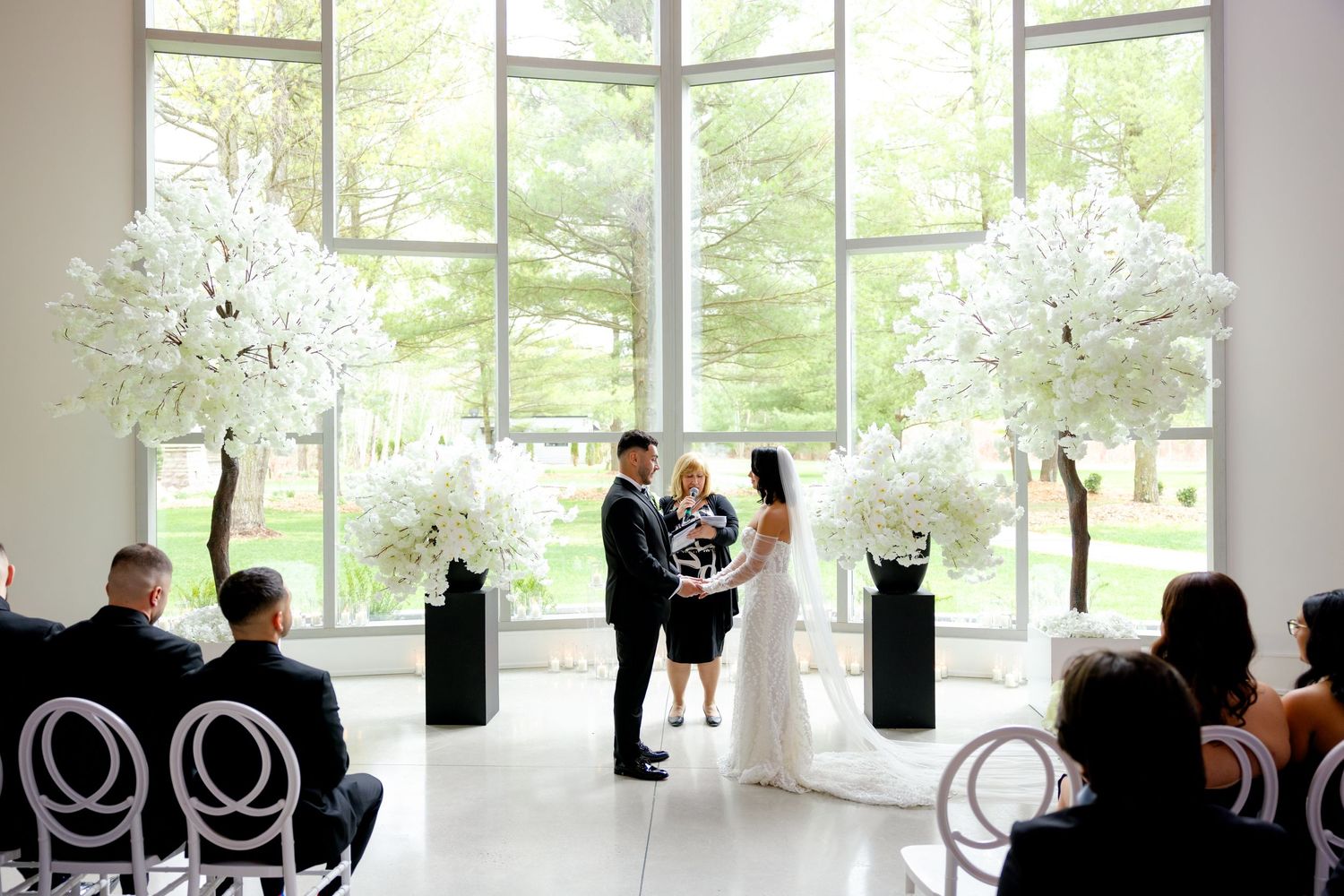 Wedding ceremony in modern venue with floor-to-ceiling windows and white floral decorations.