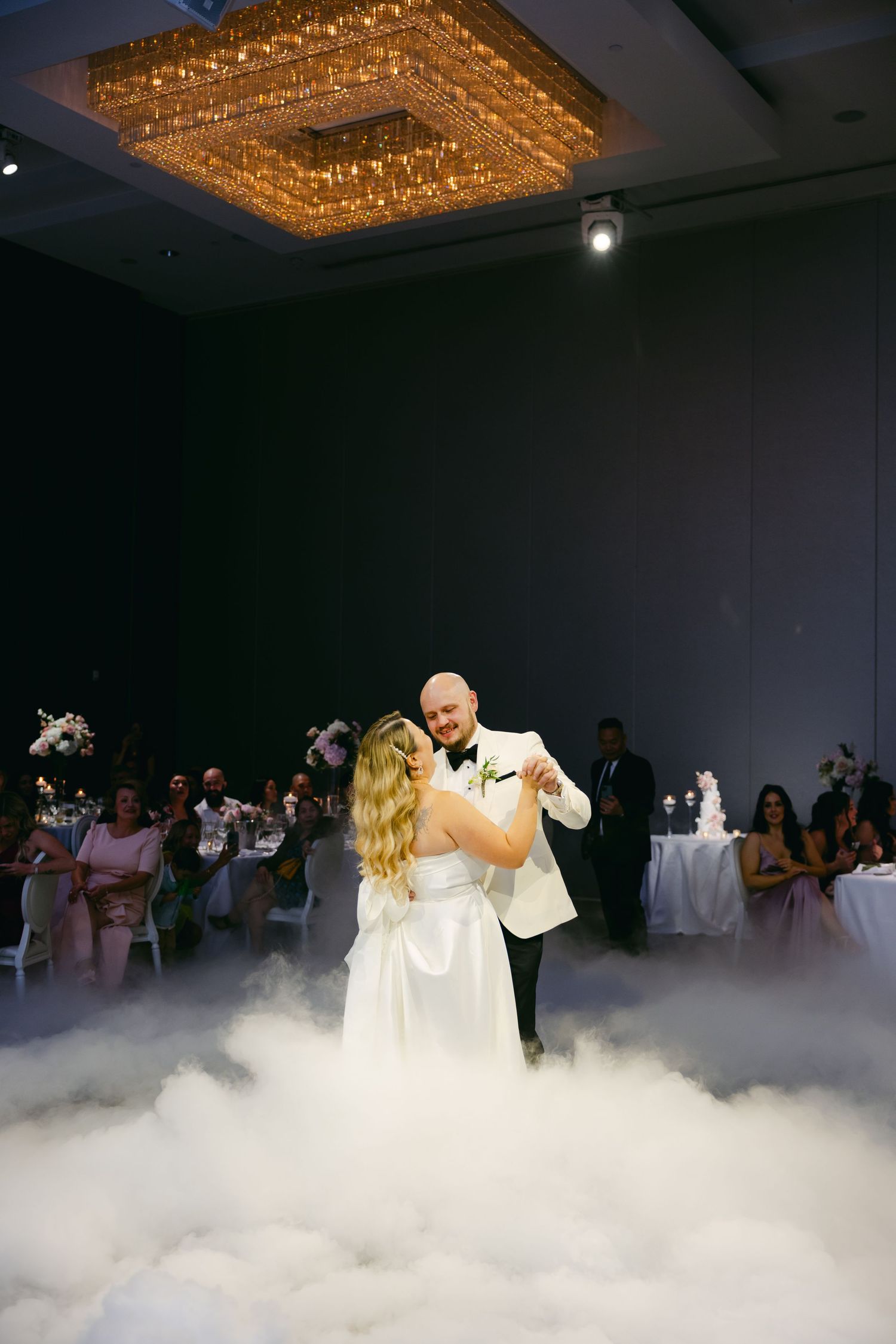 First dance moment captured under crystal chandelier with dramatic floor fog effects.