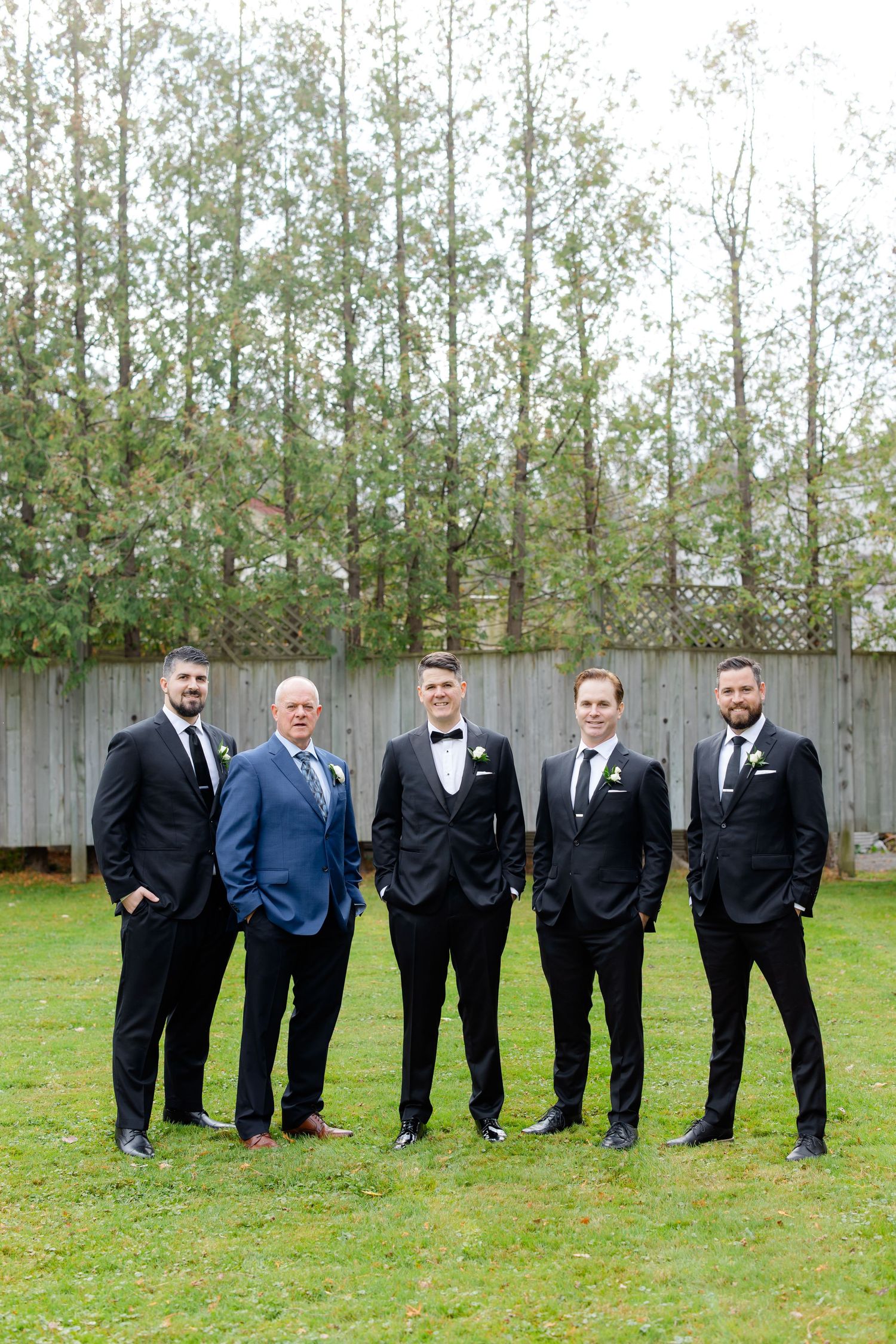 A group of five men in formal suits stand together in a garden with tall trees and a wooden fence.