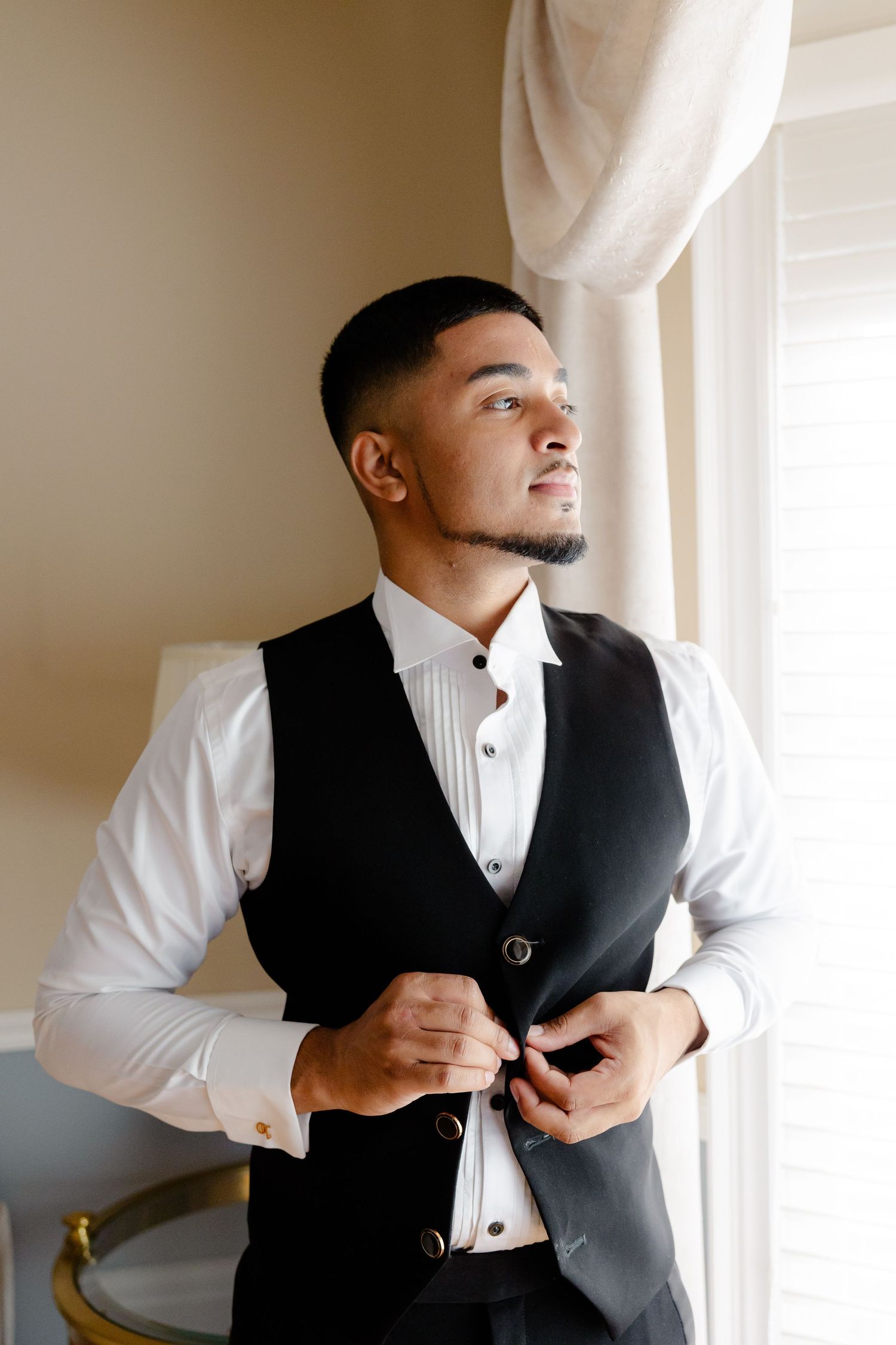 A person adjusts their formal black vest while wearing a crisp white shirt in natural window light.