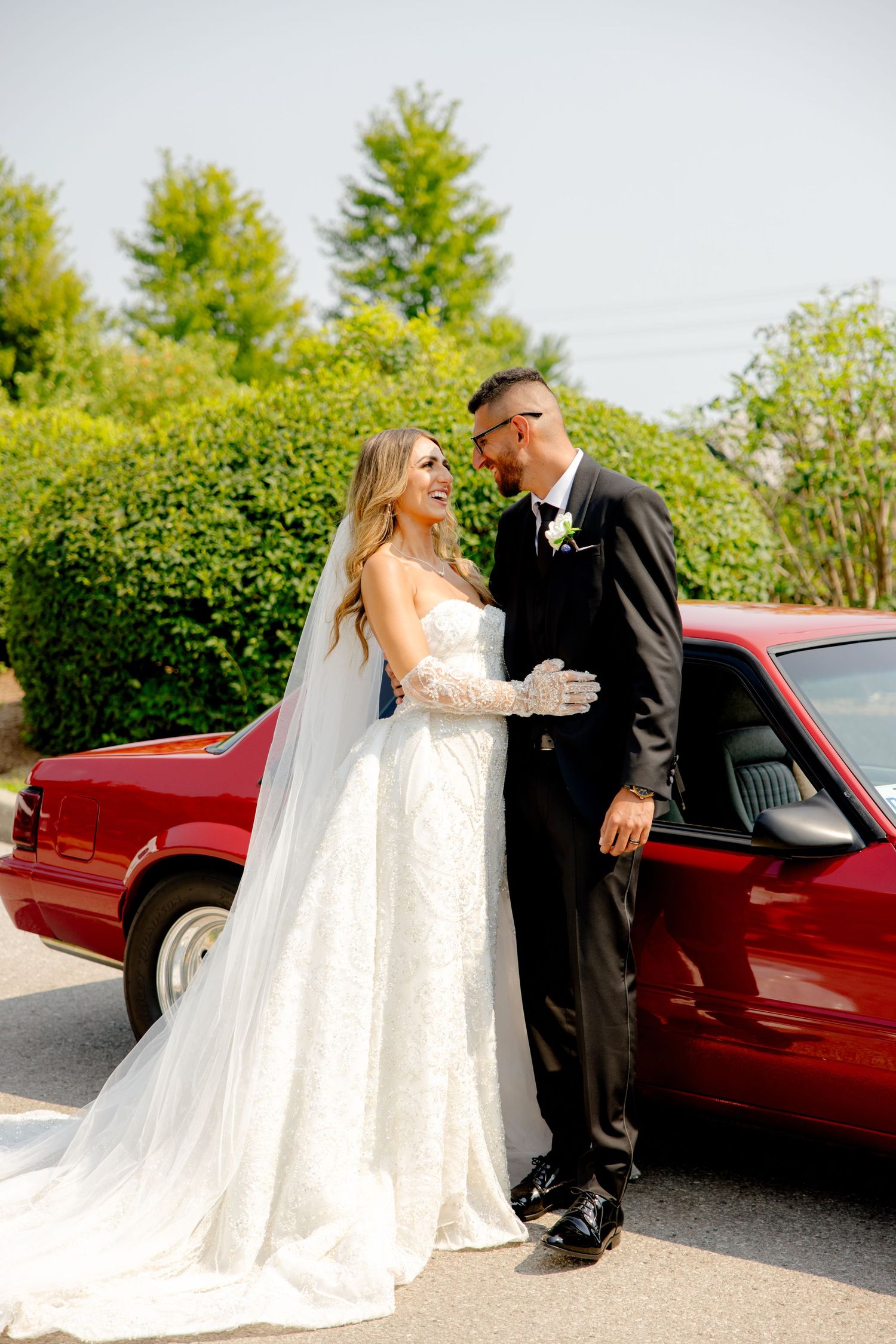 Romantic wedding photo with couple embracing beside red classic car in garden setting.