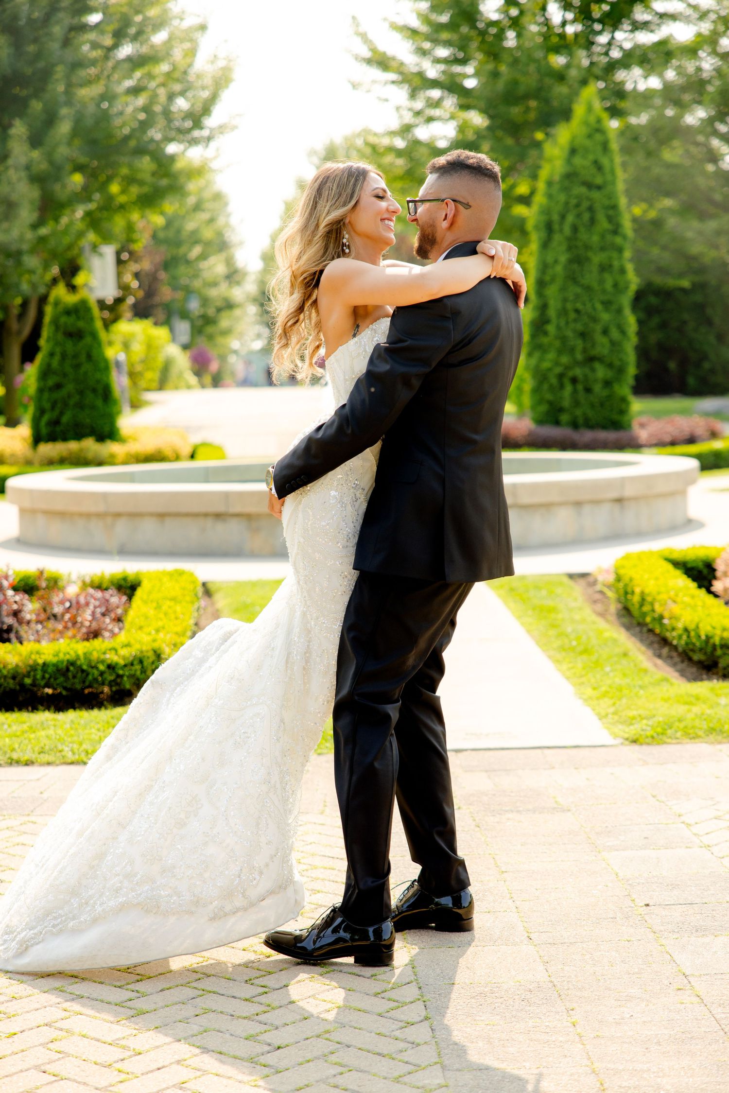 Romantic dance pose between newlyweds on brick pathway surrounded by manicured garden and fountain.