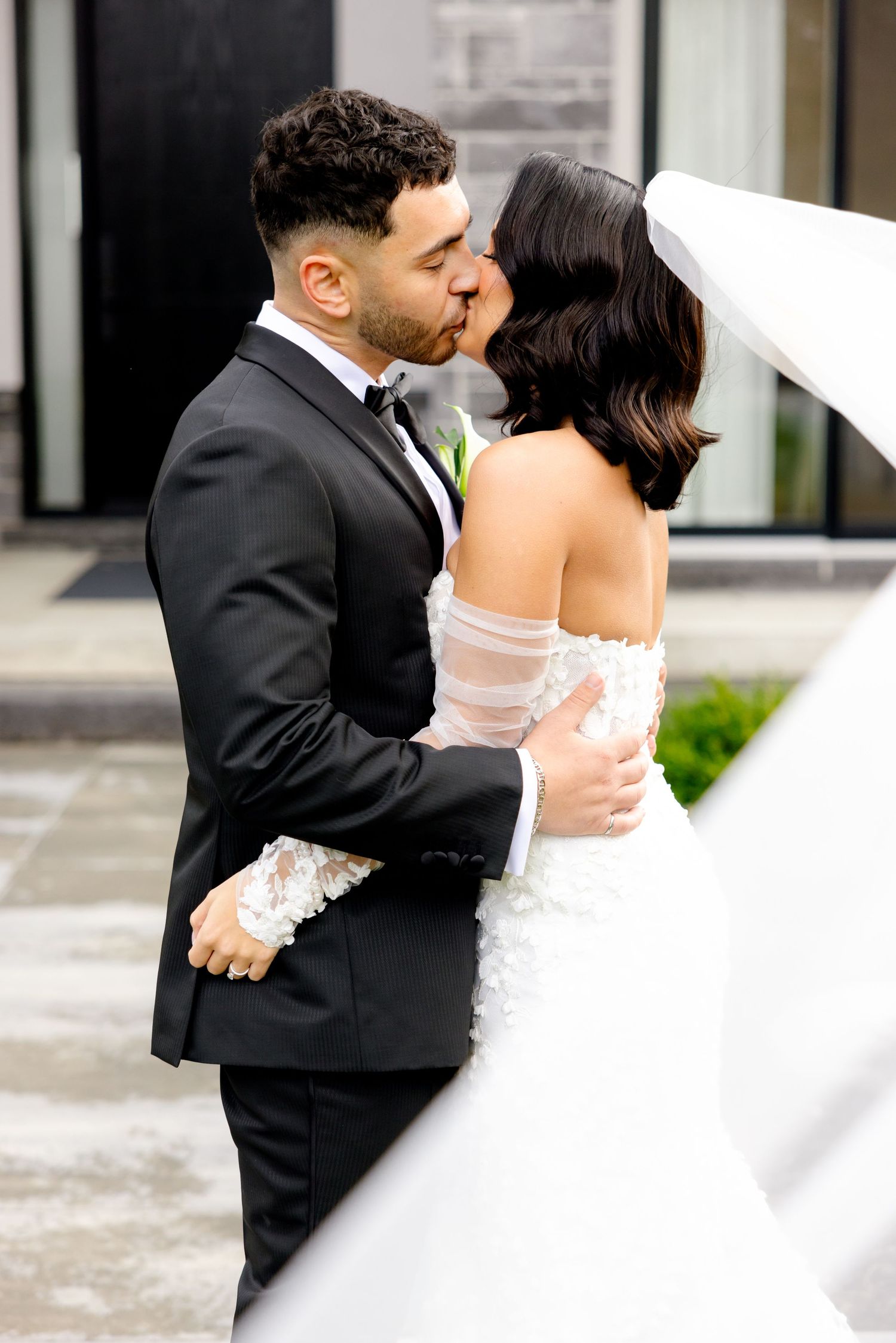Intimate wedding moment captured as couple embraces in front of stone building with flowing white veil.