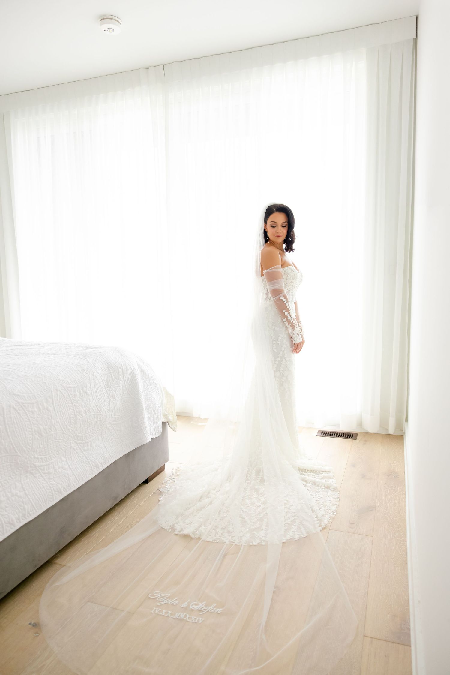 Full-length portrait of a bride in a fitted lace gown standing against white curtains.