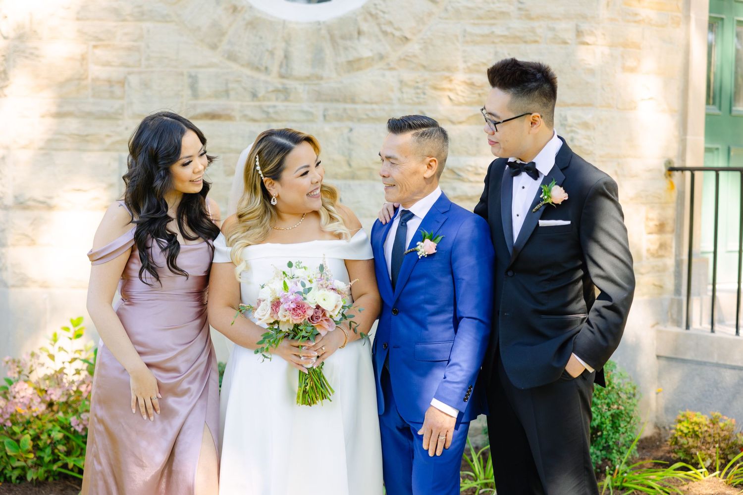 Wedding party of four shares candid moment against stone wall backdrop.