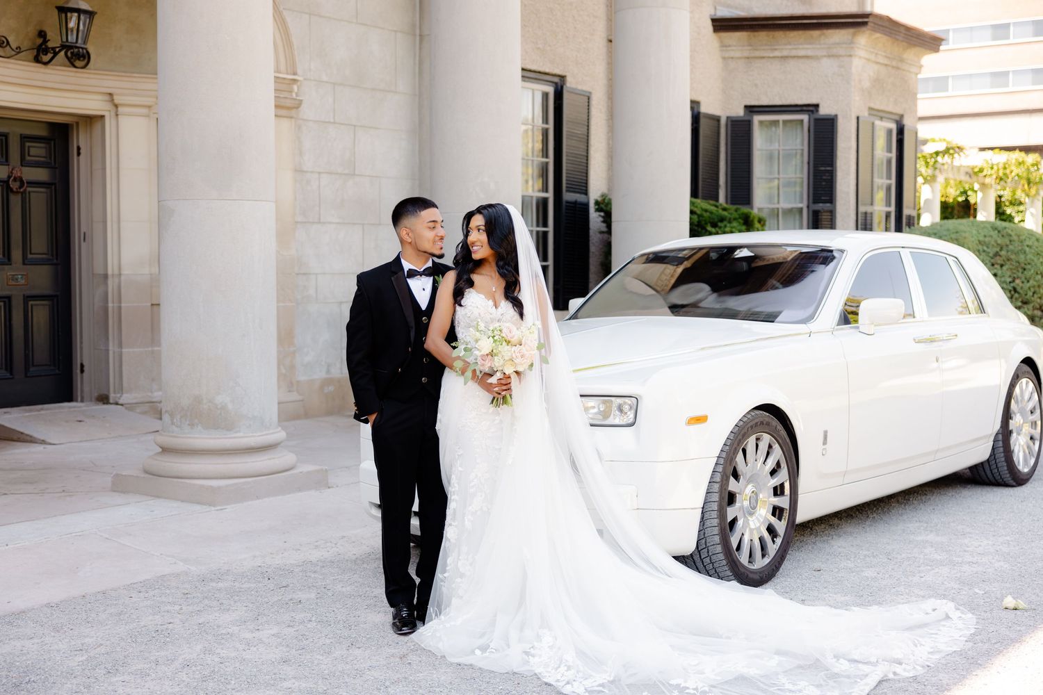 Newlyweds pose together near white luxury vehicle outside elegant building with columns.