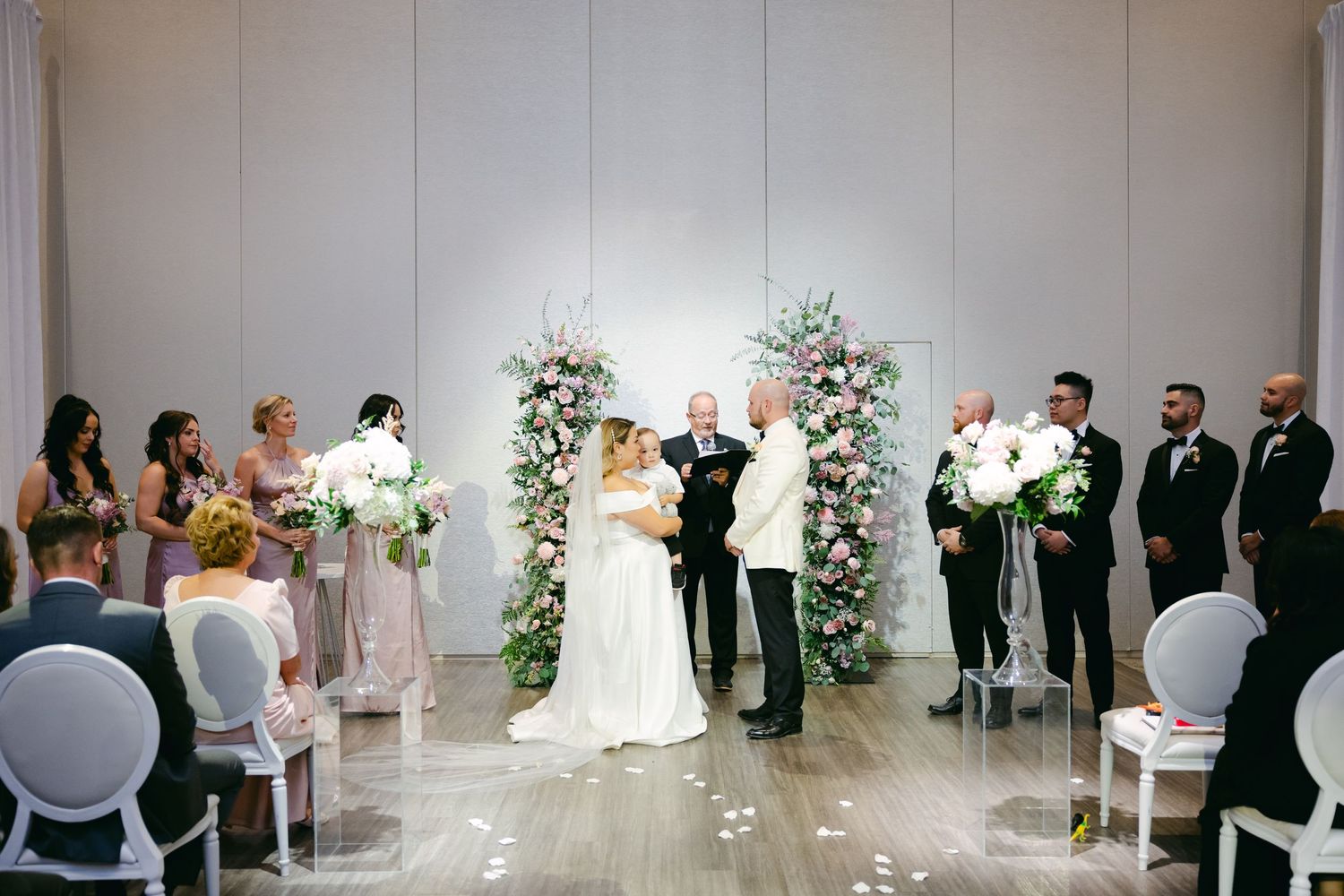 Modern indoor wedding ceremony with floral arch and white chairs.