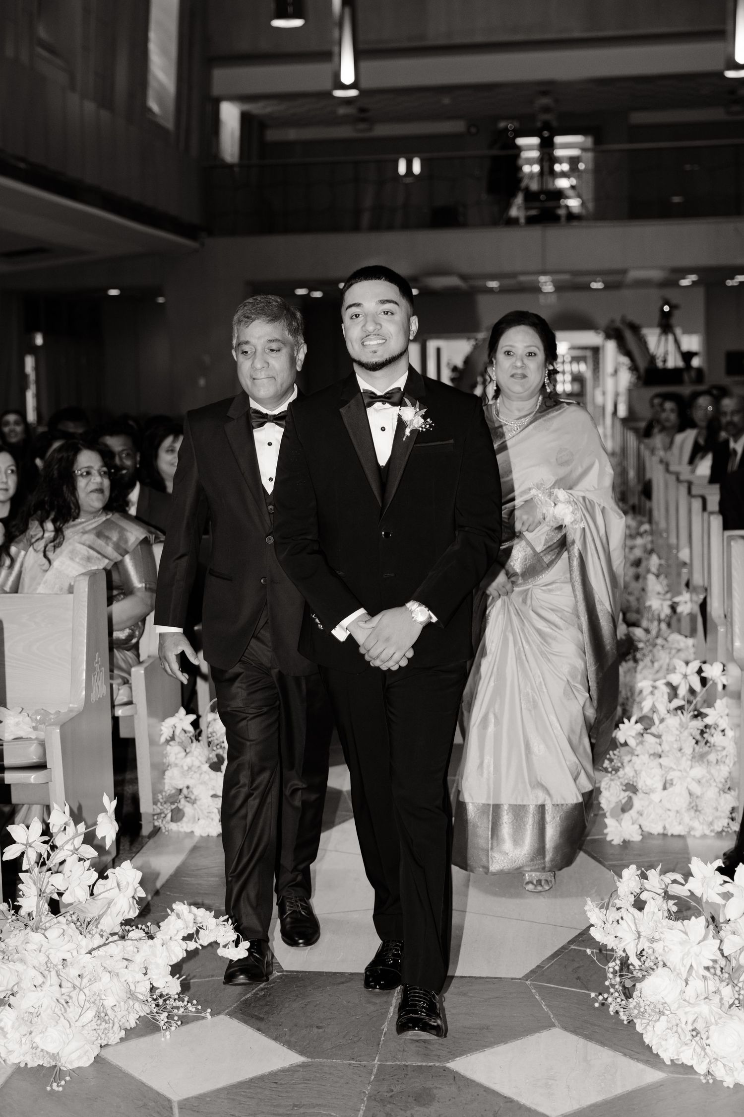 Black and white photo of wedding processional walking down decorated church aisle.