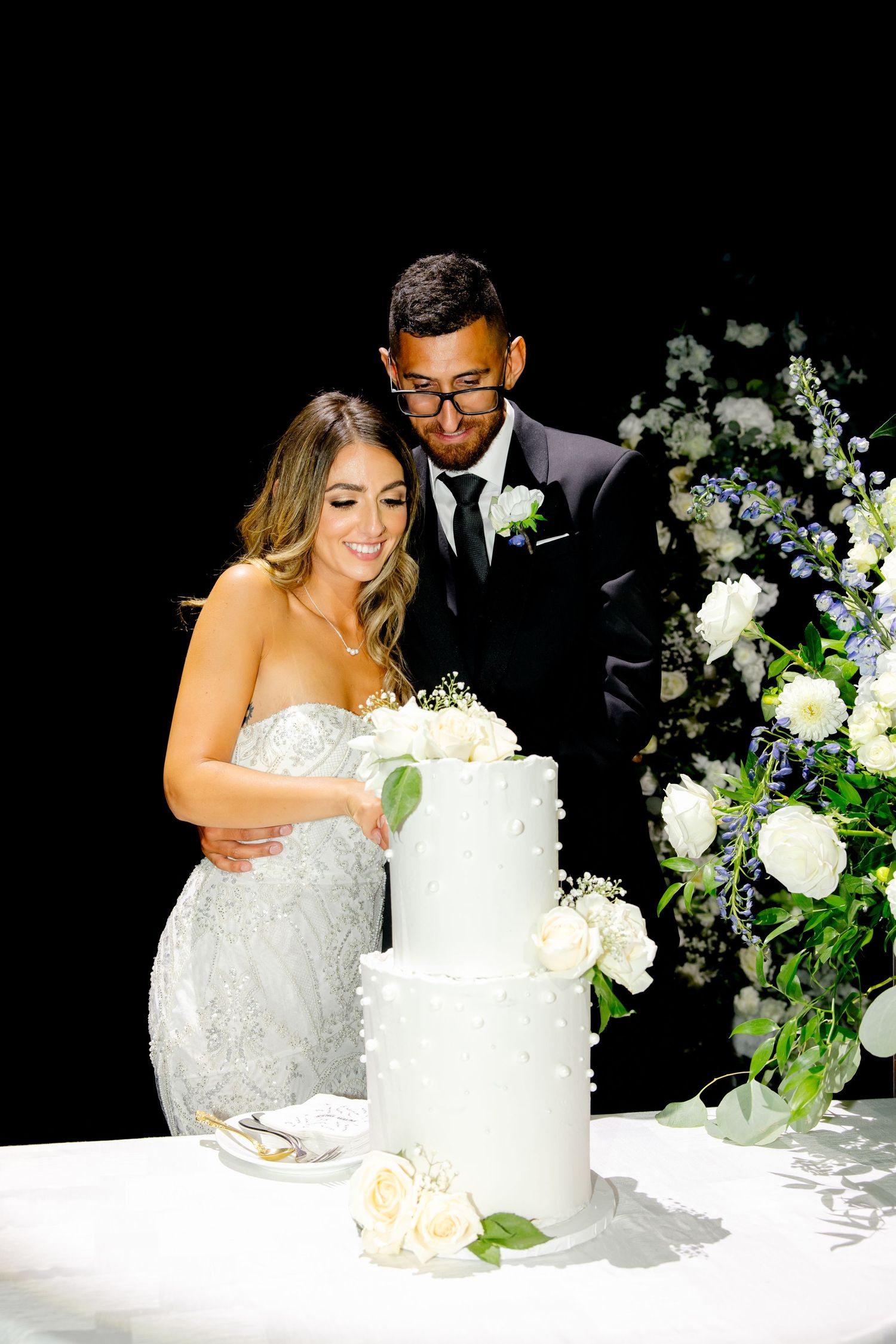 Couple cuts into an elegant white tiered wedding cake adorned with fresh flowers.