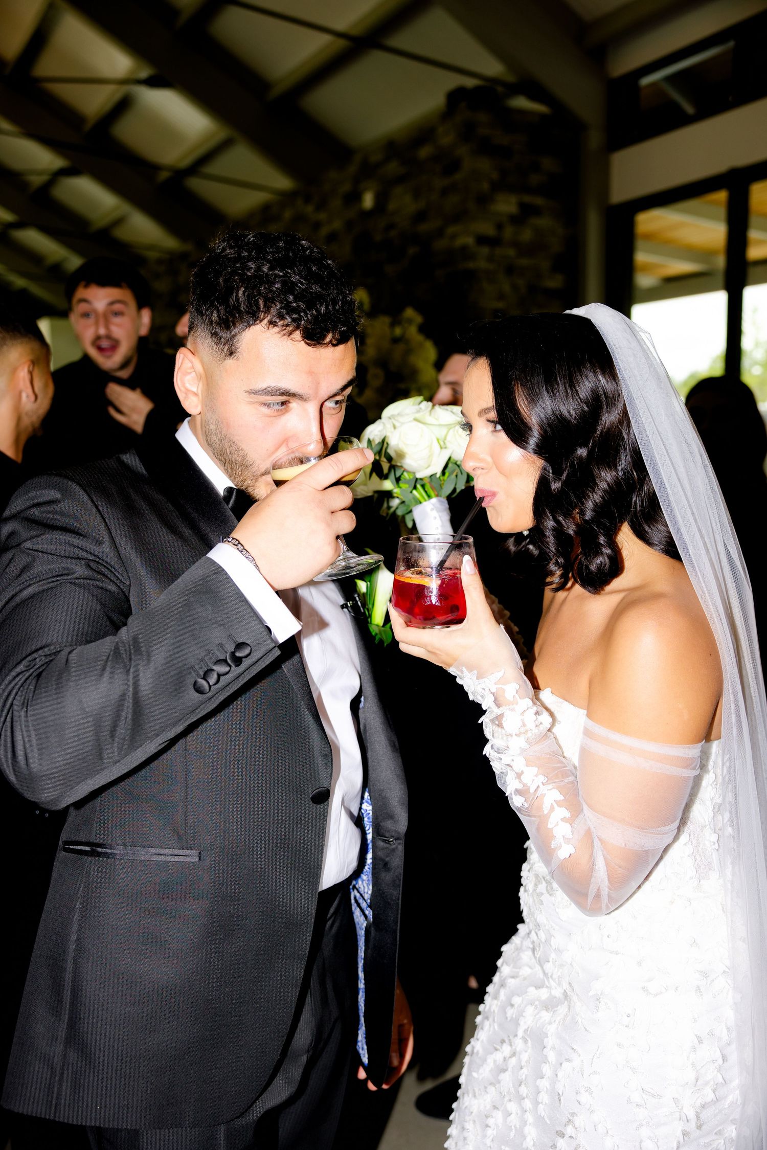 A couple shares a playful moment drinking from their wedding cocktails together.